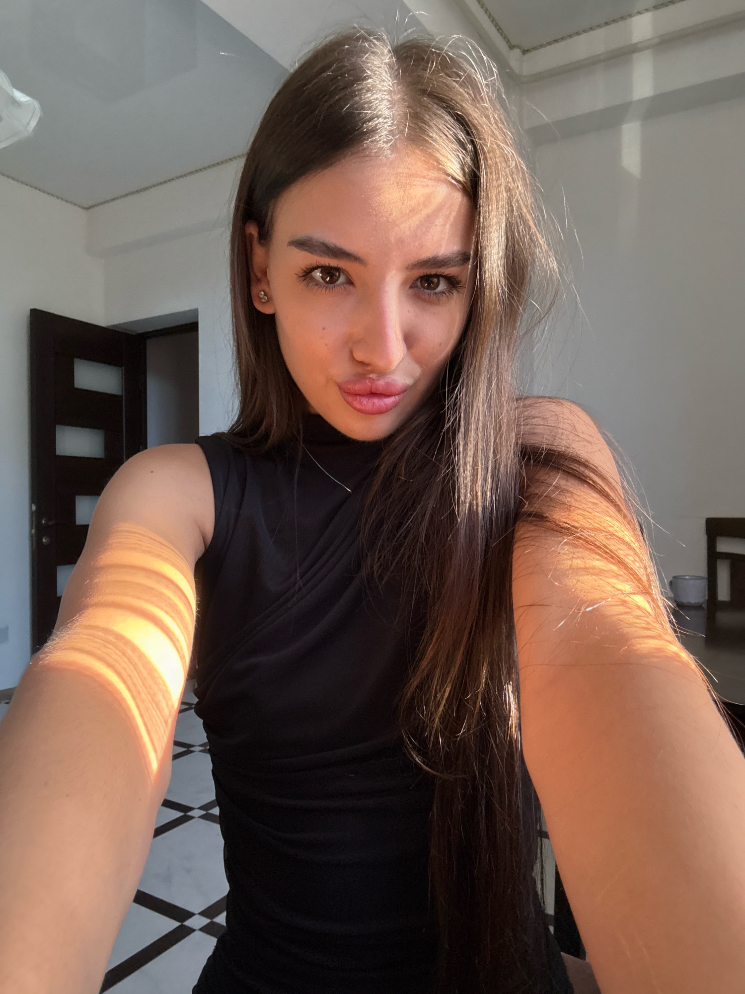 A young woman with long, dark brown hair and light skin is taking a selfie indoors. She is wearing a black, sleeveless top with a high neckline. Her head is tilted slightly, and she is puckering her lips. Sunlight streams in from the left side of the image, casting strong diagonal shadows across her face, arms, and the floor. The floor has a black and white checkered pattern. In the background, a dark wooden door with horizontal frosted glass panels is visible, along with a white wall and a portion of a chair and table on the right. There is no discernible information in the image to indicate the location in the city of Chișinău, Moldova.