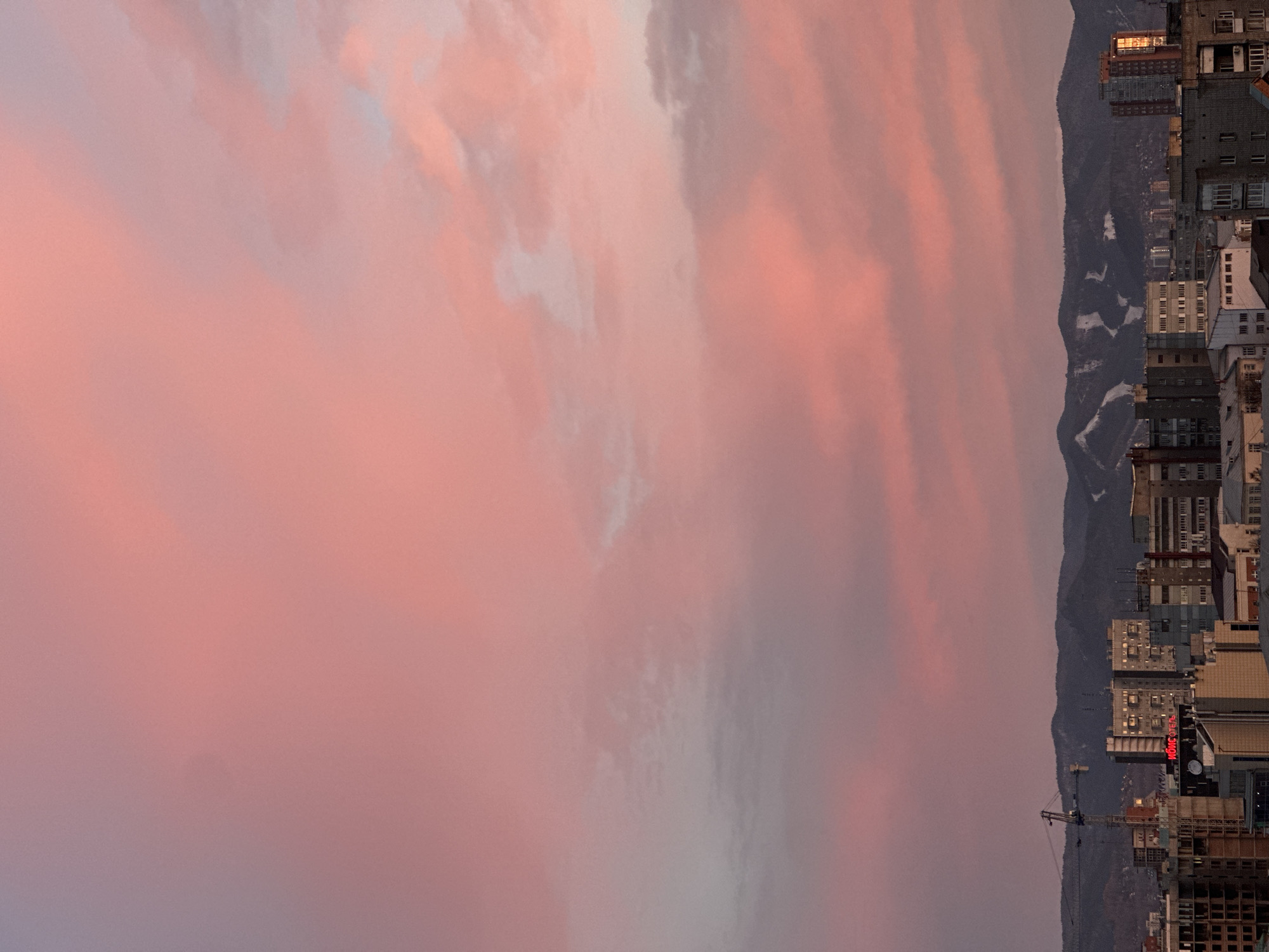 The image, taken in Krasnoyarsk, Russia, depicts an urban landscape framed by mountains under a vibrant sky, likely during either sunrise or sunset. The upper half of the frame is dominated by a dramatic sky filled with soft, diffused clouds in hues of pink, orange, and light grey.
Below the colorful sky, a range of dark, rugged mountains stretches horizontally across the middle ground. Patches of white snow are visible on the slopes and peaks, indicating either a cold climate or higher elevations.
The lower portion of the image reveals a dense cityscape nestled at the base of the mountains. Numerous multi-story buildings, varying in height and architectural style, fill the urban sprawl. Many windows are visible, some illuminated with warm interior lights, hinting at evening or early morning. A tall construction crane stands prominently on the left side of the cityscape, suggesting ongoing development.
A notable detail is the illuminated red sign on one of the buildings, displaying Cyrillic text that reads "НОМО ОТЕЛЬ" (NOMO HOTEL). Below this, another partially visible sign appears to say "БАНК" (Bank). The overall atmosphere is one of serene beauty, with the striking natural phenomenon of the sky contrasting with the structured urban environment.