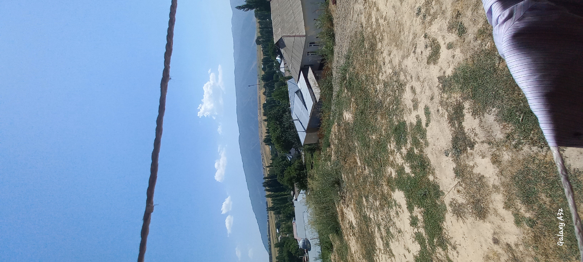 The image depicts a rural landscape under a clear blue sky. In the foreground, a rough rope runs horizontally across the frame, partially obscuring the view. Below the rope, a patch of dry, sandy ground is sparsely covered with green vegetation. Beyond this, a collection of buildings with corrugated metal and tiled roofs are visible, suggesting a small settlement. Tall, slender trees are interspersed among the houses, and further in the distance, rolling hills or mountains form the backdrop. The overall impression is of a quiet, sun-drenched day in a village, possibly located in the mountainous terrain of Joon‑Kunggöy, Kyrgyzstan. The presence of the rope and the fabric of a garment at the bottom right corner suggest the photo was taken from an elevated position, possibly a balcony or window, with laundry hanging nearby.