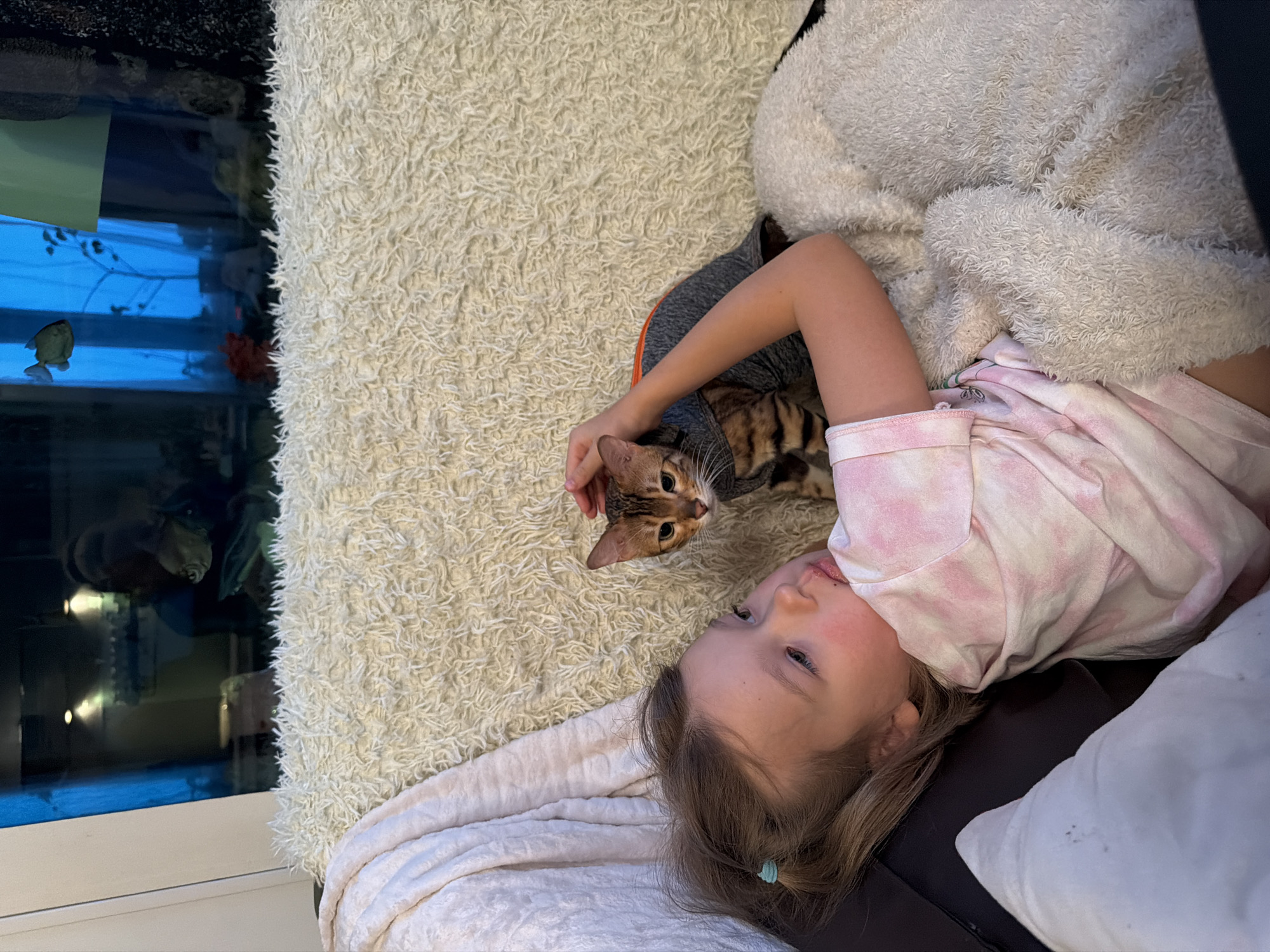 A young girl is lying on a shaggy, light-colored rug next to a Bengal cat wearing a gray and orange sweater. The girl has fair skin, blonde hair pulled back with a blue hair tie, and is wearing a pink tie-dye shirt. She is partially supported by a white pillow and a dark-colored couch. Her right hand is gently resting on the cat's head. Beside the rug is a fish tank filled with water, plants, and fish. The reflection in the glass suggests the scene is inside, possibly at night. There is no information available to identify the specific location in Saint Petersburg, Russia.