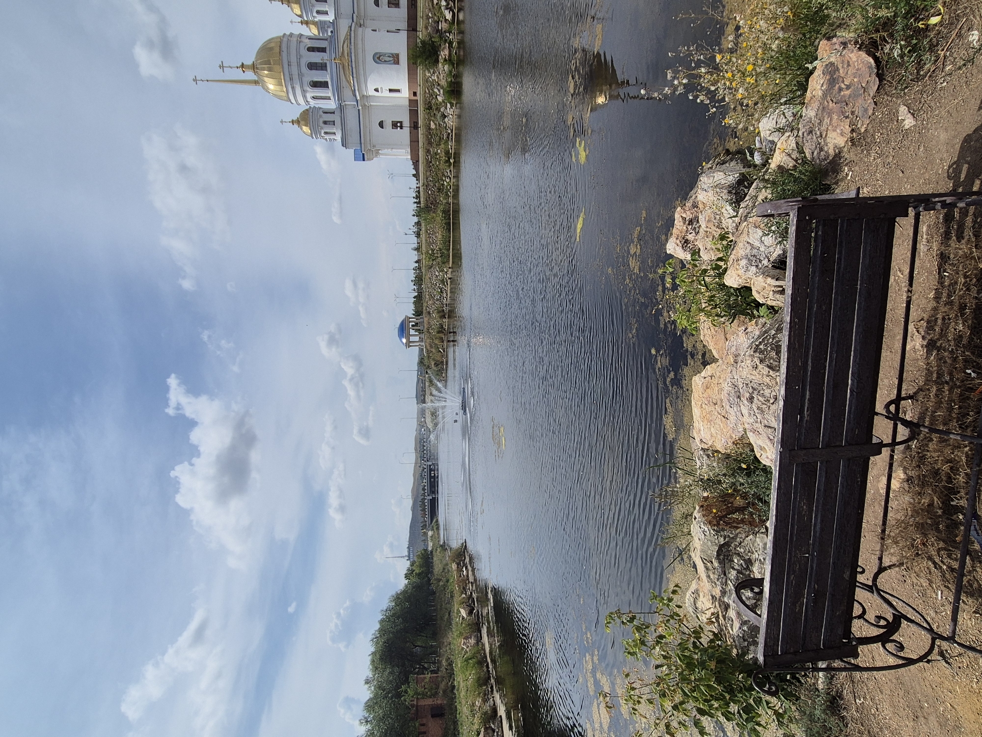 The image depicts a serene landscape with a body of water in the foreground, featuring a fountain spraying water. On the right side of the water, a white cathedral with golden domes is prominently visible, reflecting in the water. The left side of the water has a tree line and a small brick structure. In the middle ground, a bridge crosses the water, and a decorative blue dome structure is situated on the bank. The foreground shows a dark wooden bench situated on a dirt path with rocks and vegetation. The sky is partly cloudy with blue patches. This scene is the area near the Assumption Cathedral in Kokshetau, Kazakhstan.