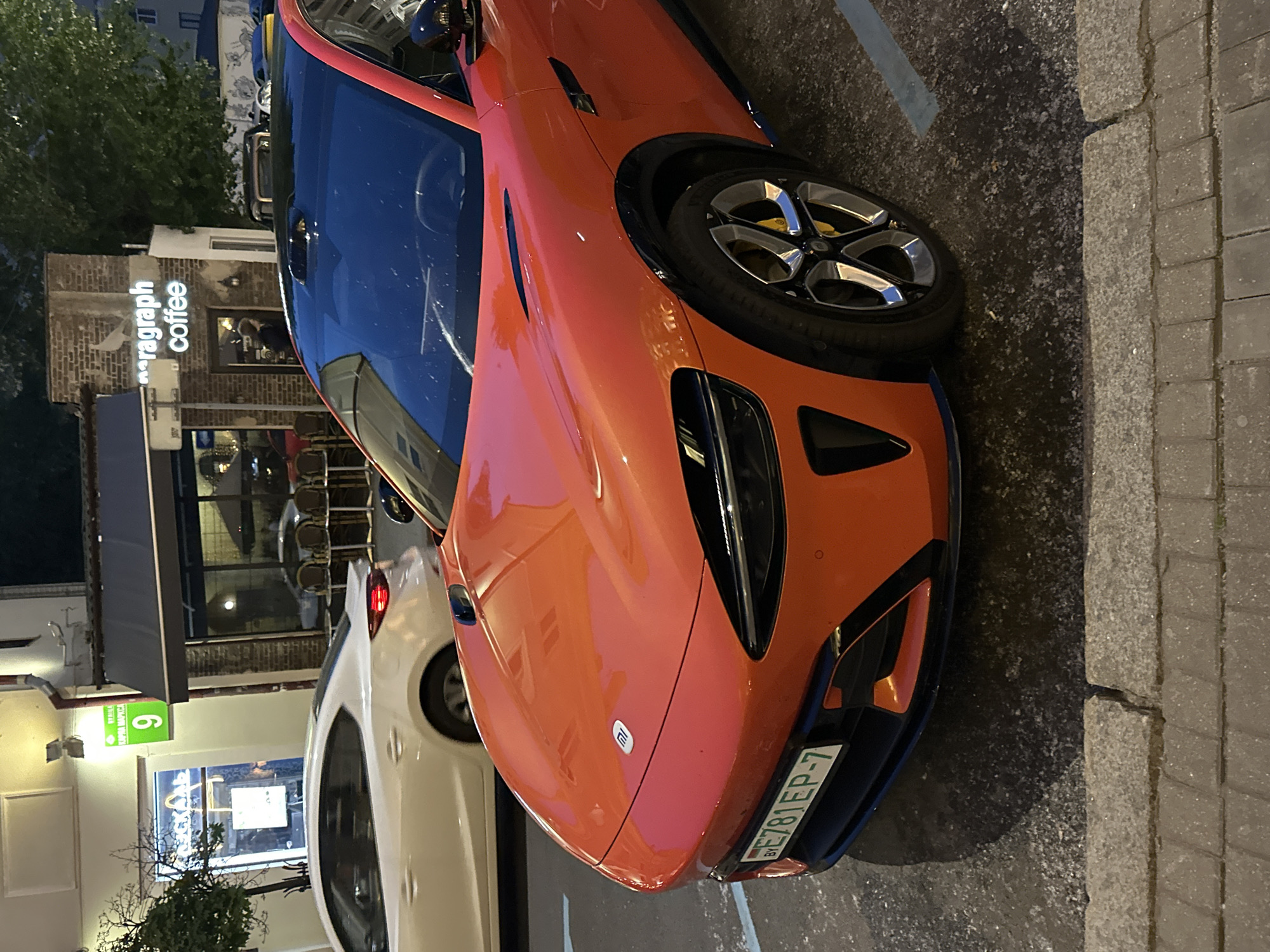 An orange Xiaomi SU7 electric vehicle, identified by its "Mi" logo on the hood and sleek design, is parked on the right side of a paved street at dusk or night. The car displays Belarusian license plate BY E781EP-7 on its front bumper. It features a black roof, prominent front air intakes with orange accents, black wheel arches, and multi-spoke wheels with visible yellow brake calipers.
In the mid-ground, a white sedan with illuminated red taillights is partially visible behind the orange car. The background reveals a street scene along Karl Marx Street 9 in Minsk, Belarus. On the left, a building with a brightly lit window displaying "ИСКАЛЬ" (ISKAL) is visible, below a green street sign indicating "КАРЛА МАРКСА 9." To the right, a brick building houses "paragraph coffee," identifiable by its illuminated sign. This café features outdoor seating with chairs and tables under a dark awning. A single person is vaguely discernible within the café's interior. The environment includes trees on both sides of the street, a paved road, and a sidewalk constructed of square pavers.