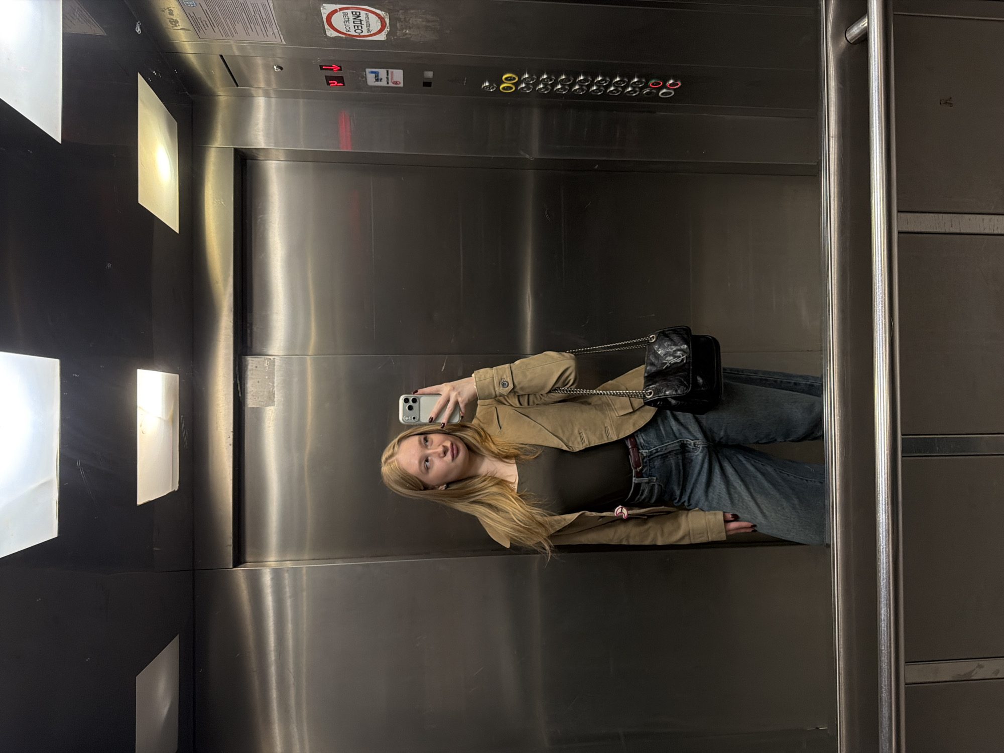A young woman stands in an elevator, taking a mirror selfie with her smartphone. She is dressed in a brown jacket, olive green shirt, and jeans, with a black purse hanging on a chain. The elevator has a metallic interior with a panel of buttons and a call button labeled in Russian. The location is likely Засечное, Russia, based on information from the image metadata or related context. The woman's action is taking a photograph, and the environment is the interior of an elevator.