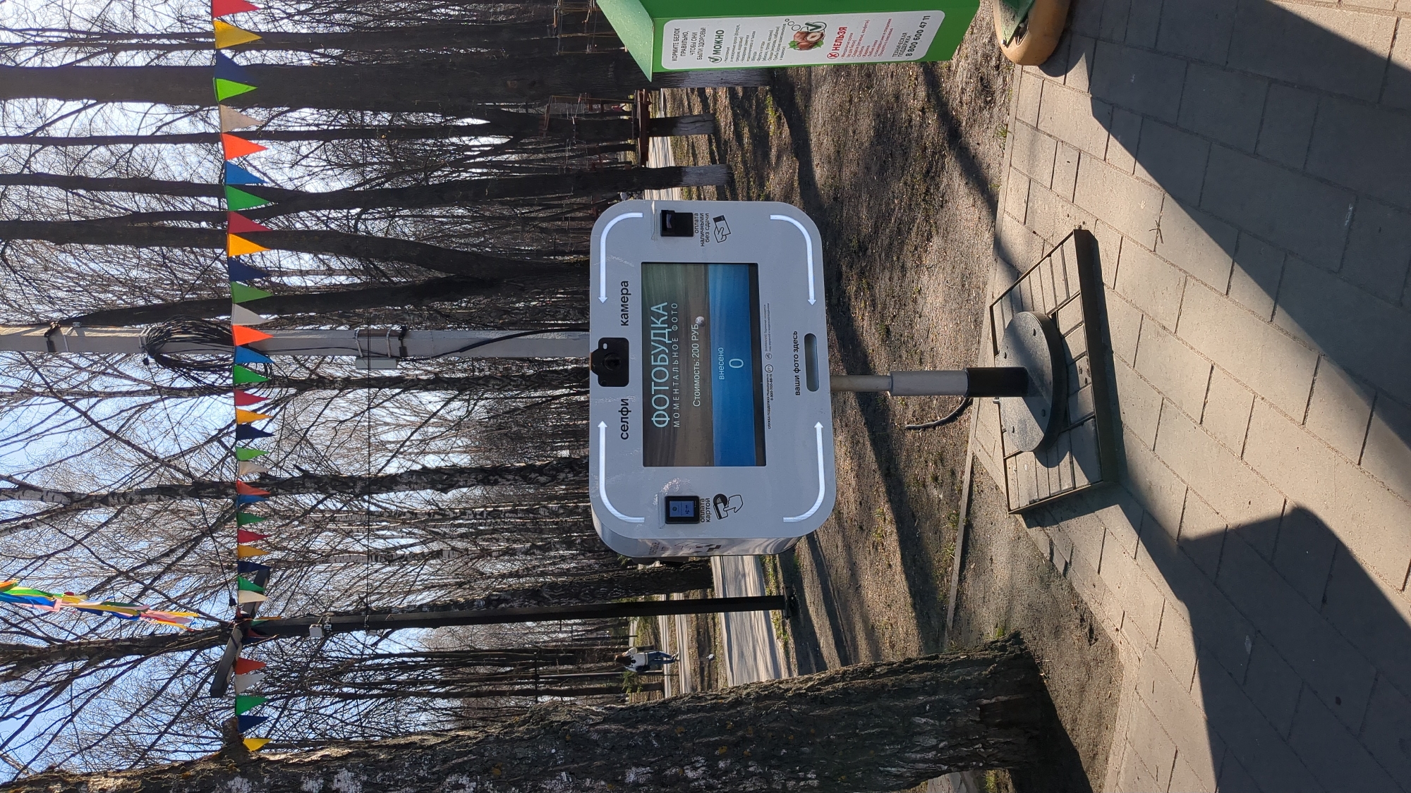 An outdoor scene in Ulyanovsk, Russia, features a self-service photobooth on a paved walkway, likely in a park or pedestrian area. The image is taken on a sunny day, with long shadows cast across the interlocking grey pavers, suggesting either early morning or late afternoon.
The main subject is a light grey photobooth, labeled "ФОТОБУДКА" (PHOTOBOOTH), which offers "МОМЕНТАЛЬНОЕ ФОТО" (INSTANT PHOTO) for "Стоимость: 200 РУБ" (Cost: 200 RUB). Its screen indicates "внесено 0" (deposited 0), implying it's ready for use. Payment options include "оплата картой" (pay by card) and "оплата наличными без сдачи" (cash payment without change). Text on the booth also points to "селфи" (selfie), "камера" (camera), and "ваши фото здесь" (your photos here) for photo retrieval.
To the right of the photobooth is a green bin, possibly for animal feed or waste, with instructions in Russian: "КОРМИТЕ БЕЛОК ПРАВИЛЬНО, ЧТОБЫ ОНИ БЫЛИ ЗДОРОВЫ" (FEED SQUIRRELS CORRECTLY SO THEY ARE HEALTHY), followed by categories "МОЖНО" (CAN) and "НЕЛЬЗЯ" (CANNOT), and a "ТЕХНИЧЕСКАЯ ПОДДЕРЖКА" (TECHNICAL SUPPORT) phone number: "8 800 600 47 11".
Bare-branched trees line the path, indicating late winter or early spring. Colorful triangular flags are strung between the trees, adding a decorative or festive touch to the park environment. In the distance, a person can be faintly seen walking on a path through the trees. The sky is clear and blue.