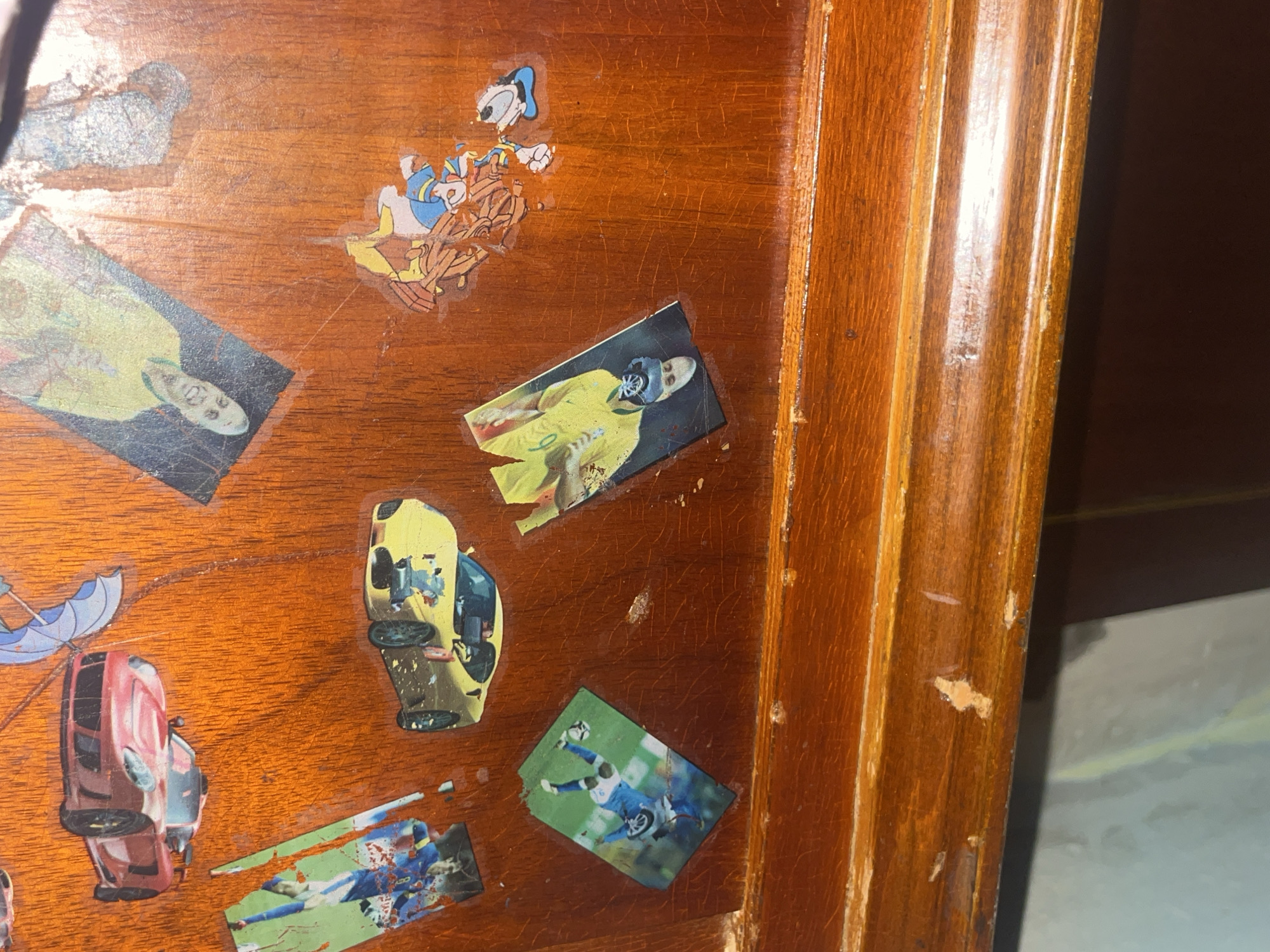 The image shows a close-up view of a wooden surface, possibly a desk or a table, adorned with several stickers. The surface is a warm, reddish-brown color and exhibits visible wood grain and some wear and tear, including scratches and chipped paint. On the right side of the image, a vertical wooden trim with fluted detailing is visible.
The stickers depict various subjects. In the upper left quadrant, there's a sticker featuring Donald Duck from Disney, seemingly riding in a cart. To the right of Donald Duck, there is a sticker of what appears to be a sports trading card, showing a person in a yellow uniform with a blue helmet. Another sticker, located slightly below and to the right of the first sports card, also seems to be a sports-related image, with a person in a blue uniform, possibly playing soccer, with a ball at their feet.
Further down on the left, a sticker of a red car is visible, with an umbrella sticker above it. To the right of the red car, there is a sticker of a yellow sports car, and below that, another sports-related sticker, showing a person in a blue uniform. There are also other scattered stickers that are partially visible or obscured.
The lighting suggests an indoor setting, and there are no clear indications of the time of day or weather conditions. The overall impression is that of a decorated surface, likely in a personal space. No text is clearly discernible on the stickers. The context of Yerevan, Armenia, doesn't add specific visual cues to the image itself, as the content is universal.