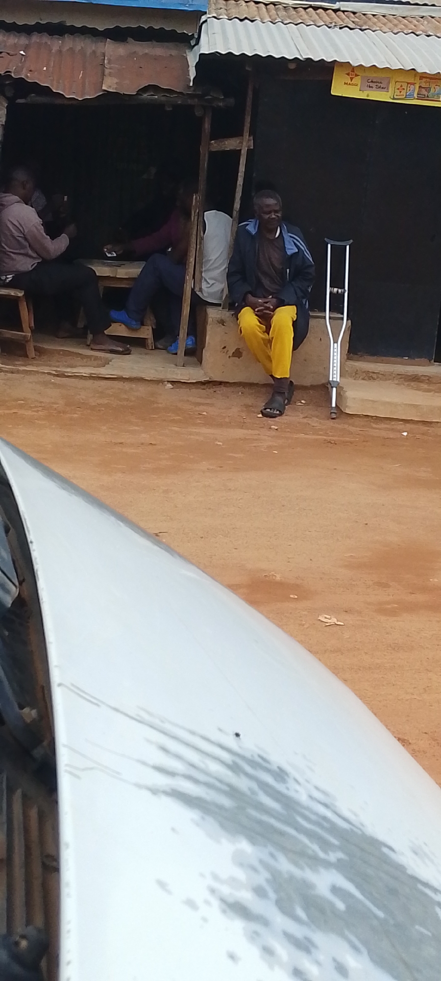 The image depicts an outdoor scene in Jos, Nigeria, likely in front of a small, makeshift shop or dwelling. The shop has a corrugated metal roof and wooden supports, with a dark interior. In the foreground, the hood of a white vehicle is visible, showing some water stains and scratches.
Within the shop area, several individuals are present. On the left, a person wearing a grey hoodie and dark pants sits on a wooden stool, holding what appears to be a phone. Next to them, another person in a purple top and dark pants is also seated. Another individual, partially obscured, is seated behind them.
To the right, a man dressed in a dark jacket with blue stripes over a brown shirt and bright yellow pants is seated on a low concrete step. He is wearing dark sandals. Beside him, leaning upright against the shop structure, is a silver forearm crutch. The ground in front of the shop is reddish-brown dirt. A yellow sign with text, including the word "MAGGI," is attached to the shop's exterior. The overall impression is a casual gathering or waiting area outside a local business.