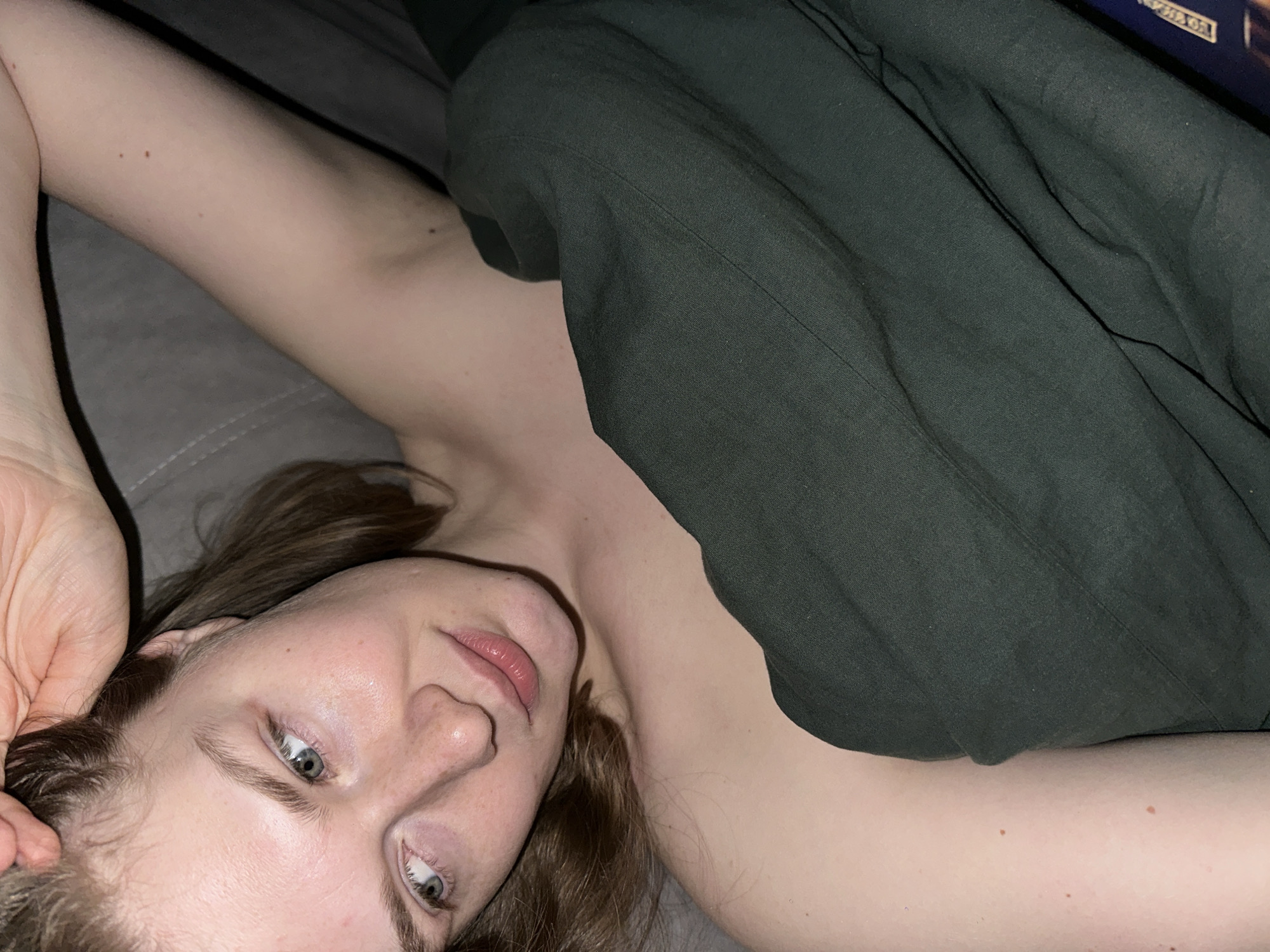 A woman with light skin, brown hair, and blue eyes is lying on a gray surface. She is partially covered by a dark green garment. One of her arms is raised with her hand near her head. The location is unknown and there is no visible indication of it being Kazan, Russia.