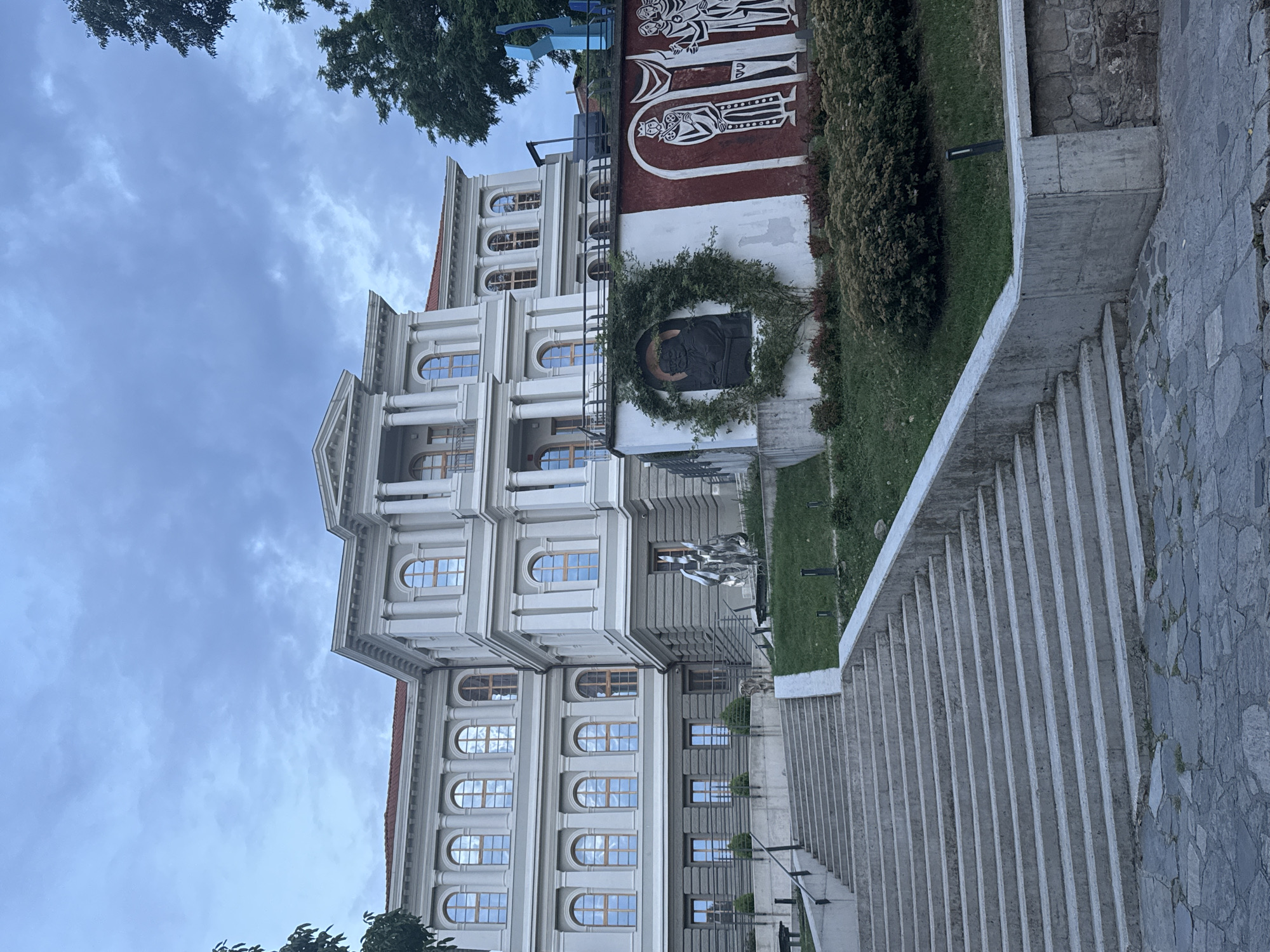 The image depicts a neoclassical building with multiple arched windows and ornate architectural details, set against a cloudy sky. In front of the building, a large wreath encircles a dark relief sculpture. Above the sculpture, a mural with stylized figures in black and white on a red background is visible. To the right of the building, a concrete staircase with a railing leads upwards, with a stone-paved path alongside. The scene appears to be an outdoor public space, possibly a museum or cultural institution, given the artwork. The lighting suggests it might be late afternoon or early morning, with overcast conditions. The presence of trees and manicured greenery indicates a well-maintained environment. There is no visible text in the image.