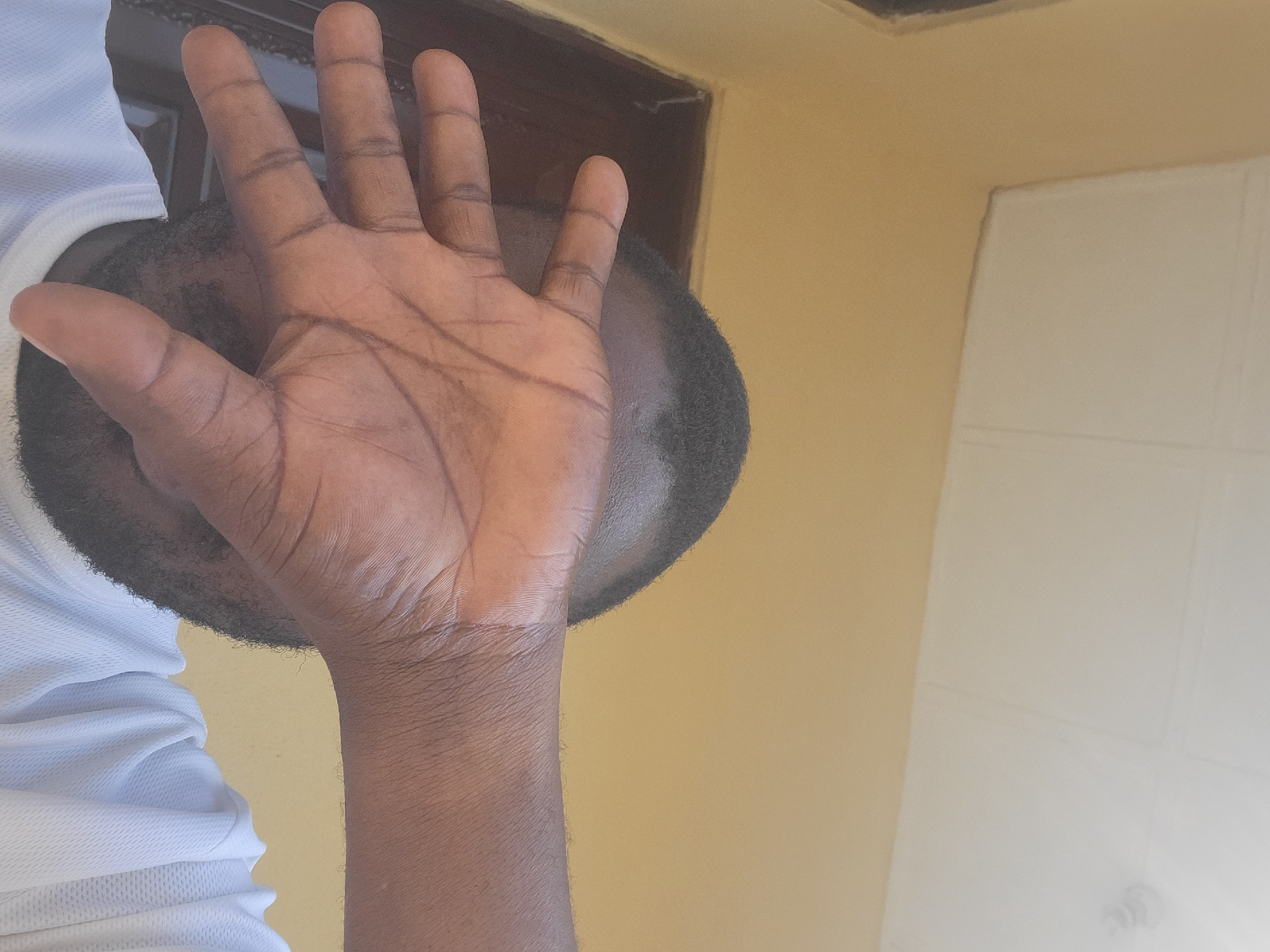 The image is a close-up, low-angle shot of a person covering their face with their right hand. The person's hand, with visible palm lines and dark skin, is positioned in front of their eyes. Part of their head and a white shirt are also visible. The background consists of a yellow wall and a white ceiling with paneling. A dark doorway is partially visible to the left. The scene appears to be indoors, likely in a residential or office setting in Suleja, Nigeria. The lighting suggests it is daytime, possibly with overhead illumination contributing to the brightness of the ceiling.