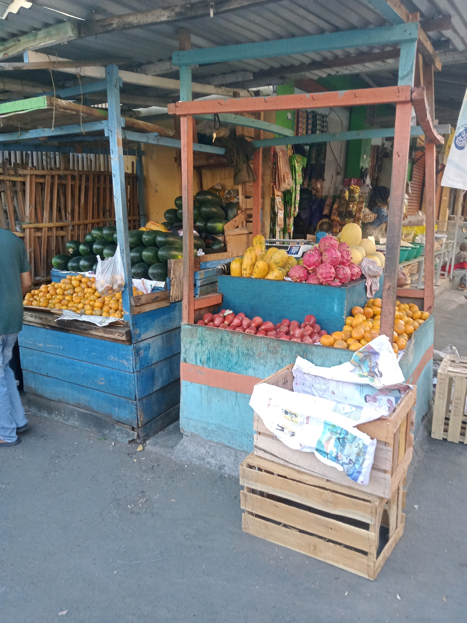A street-side fruit stall in Cirebon, Indonesia, is captured in this medium shot. The stall is constructed from weathered wood and painted in vibrant blue and orange hues. A variety of fresh fruits are displayed, including stacks of watermelons, a pile of small orange fruits resembling tangerines, and an assortment of tropical fruits like papayas and dragon fruit. Empty wooden crates are stacked in the foreground, one holding crumpled newspapers. A person wearing a green t-shirt and blue jeans is partially visible on the left, walking away from the stall. The ground is paved with asphalt, and a corrugated metal roof covers the stall, suggesting an outdoor market setting. The background shows a glimpse of other stalls and structures, indicative of a bustling marketplace.