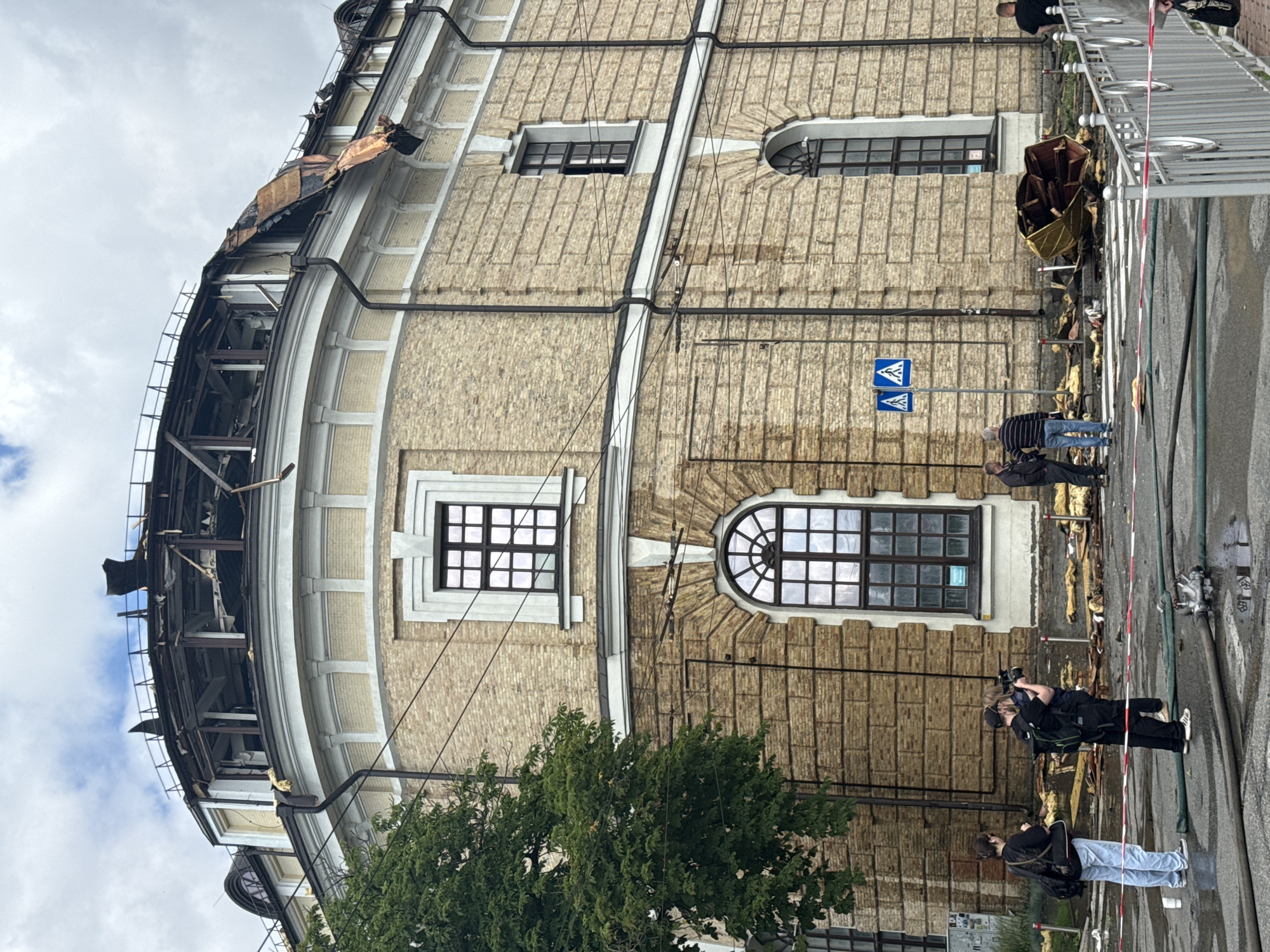 The image depicts the aftermath of an explosion or fire at a building in Kyiv, Ukraine. The top of the building, a circular structure with beige brickwork and white decorative elements, is severely damaged, with the roof structure exposed and debris scattered. Several people are present in the foreground, including individuals filming or photographing the scene, and others standing or observing. The street in front of the building is wet and shows signs of debris, including what appears to be water hoses. A pedestrian crossing is visible, marked with red and white tape. The sky is overcast.