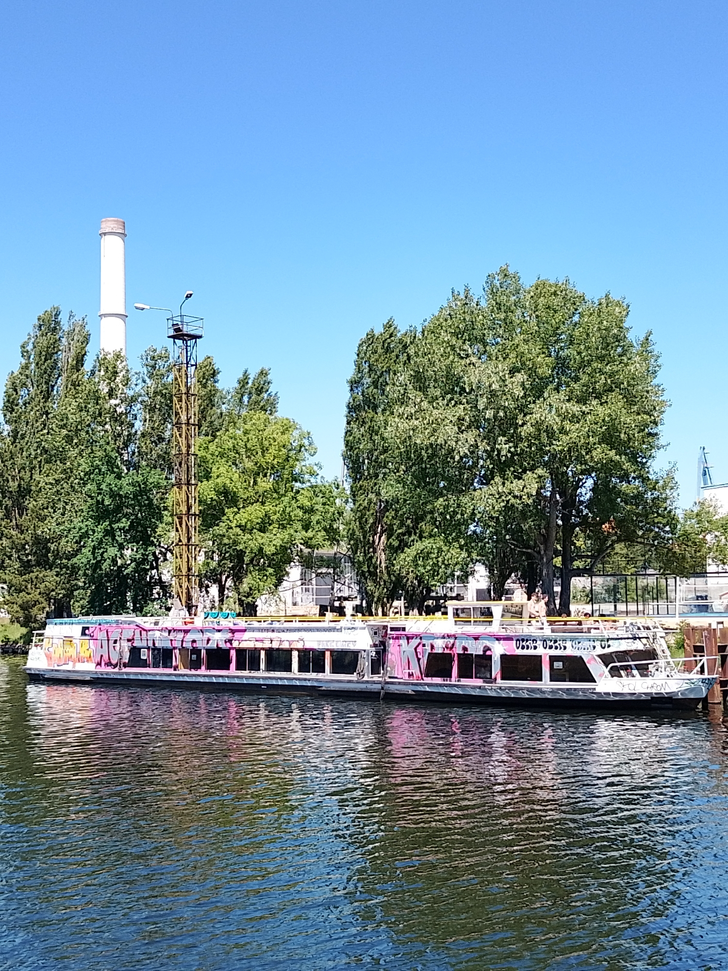 The image shows a canal scene on a sunny day. A boat with graffiti is docked alongside a pier. The boat has a silver hull and pink and white graffiti on the upper deck and sides. Its windows are rectangular and dark. Two people are standing on the upper deck. In the background are green trees and a tall, white smokestack. To the left of the trees is a narrow, lattice tower with a small platform at the top and a streetlight attached. The water in the canal is dark with reflections of the boat and sky.