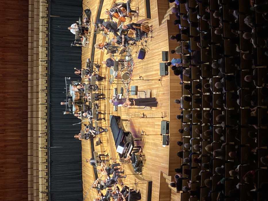 The image captures a high-angle view of a live musical performance in the Symphony Hall (Sala Sinfónica) at the Centro Cultural Kirchner (CCK) in Buenos Aires, Argentina. The large, multi-tiered wooden stage is brightly illuminated, featuring an ensemble of approximately 30-40 musicians. These musicians are seated at various levels, performing with instruments including violins, cellos, a double bass, flutes, clarinets, percussion (timpani, congas, a drum kit), and a prominent black grand piano. Music stands are positioned in front of most performers.
Centered on the lower stage is a female vocalist, dressed in a dark, embellished top and wide-leg trousers, standing with arms slightly extended, facing the unseen audience. Adjacent to her, a male conductor in a dark suit stands with his back partially to the camera. A female pianist is seated at the grand piano on the left side of the stage. Numerous microphones on stands and stage monitors are arranged across the stage floor. The stage's backdrop consists of a dark, pleated curtain, above which rises a curved, light-colored wooden wall.
In the foreground, multiple rows of tiered seating are visible, largely occupied by an audience observing the performance. The audience area is dimly lit compared to the stage.