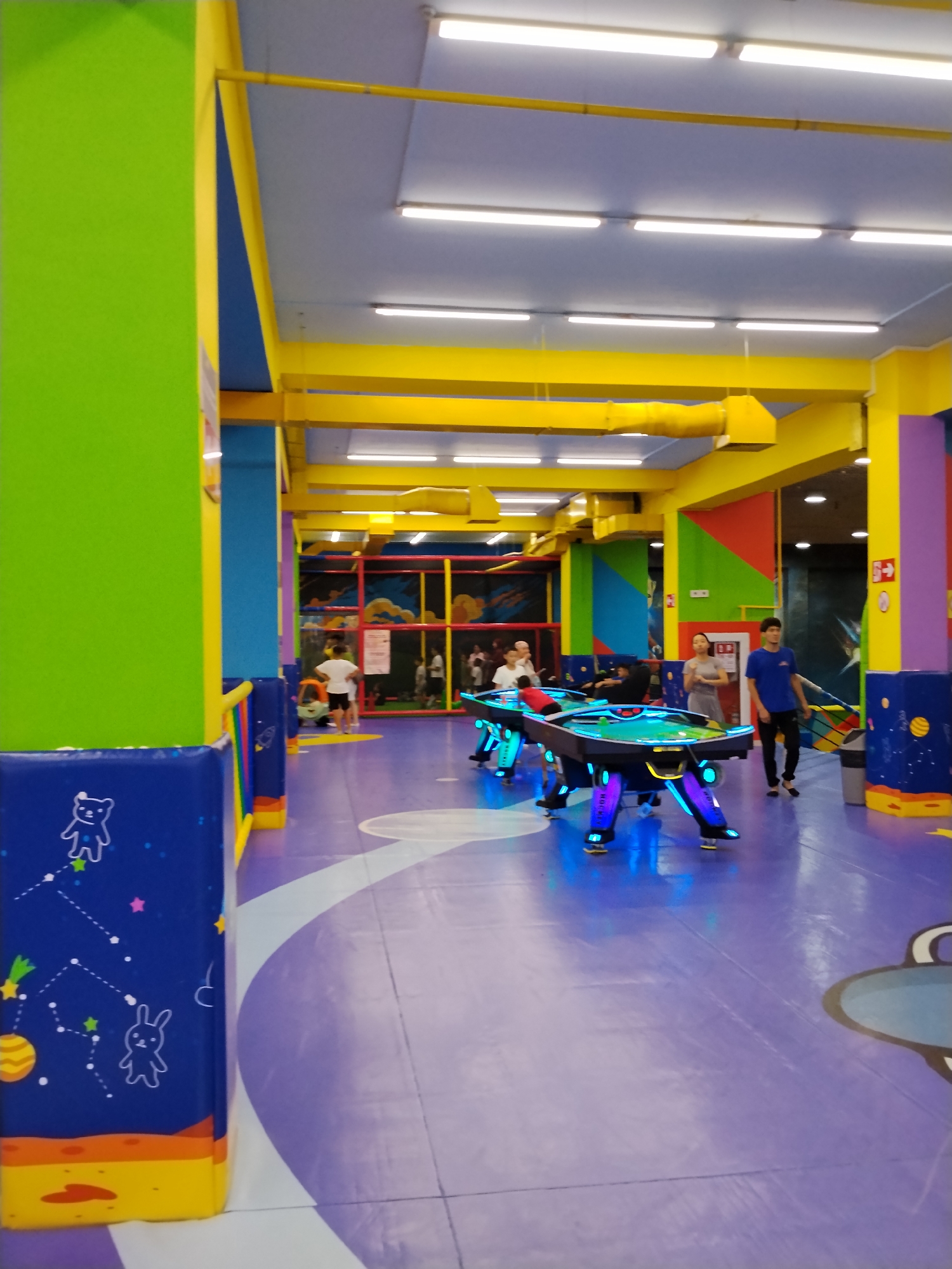 The image depicts an indoor amusement center, likely in Bishkek City, Kyrgyzstan. The environment is brightly colored with dominant hues of green, yellow, blue, and purple. A padded play area with soft barriers featuring celestial and cartoon motifs is visible on the left. In the center, a group of people are gathered around a futuristic-looking pool table with neon blue and purple lights. The table is intricately designed with illuminated legs. Several individuals, including children and adults, are present, some appearing to play or observe the game. The floor is a light purple color with white curved markings. In the background, there is a soft play structure with red, yellow, and blue netting and slides, indicating a children's play zone. The ceiling features fluorescent lighting and yellow structural beams. The overall scene suggests a lively and family-oriented entertainment venue.