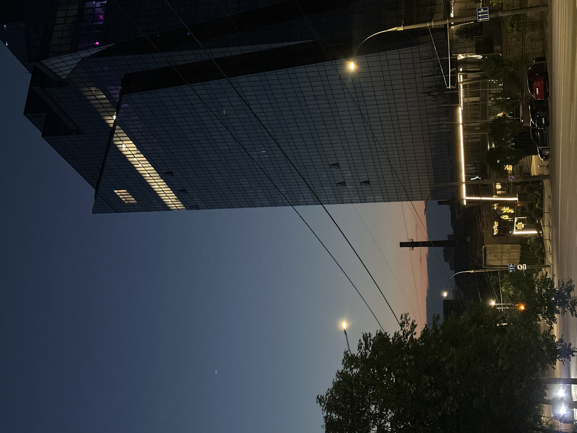 A nighttime shot of a city street in Chișinău, Moldova. A modern, multi-story glass building looms overhead, with light emanating from some of its windows. On the street level, there's a line of parked cars, streetlights, and trees along the sidewalk. The sky transitions from a dark blue to a lighter gradient.