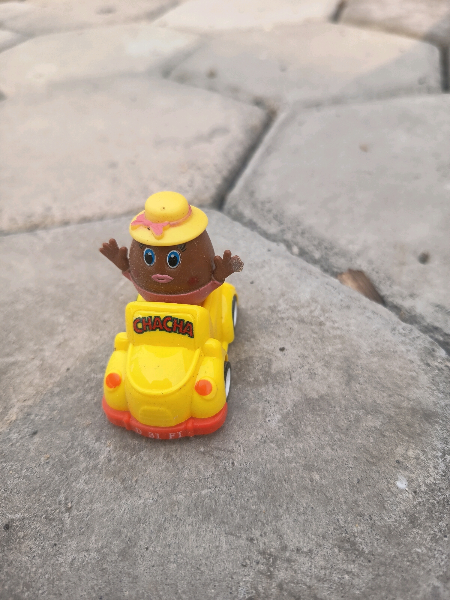 A small, yellow toy car with red trim is on a paved surface. A brown, egg-shaped character wearing a yellow hat with a pink ribbon is sitting in the car. The character has blue eyes, pink lips, and rosy cheeks. The word "CHACHA" is written in red on the back of the car. The paved surface appears to be made of large, hexagonal gray stones. In the background, the stones are out of focus, with shadows suggesting an outdoor setting. The image is taken from a low angle, focusing on the toy. This scene is located in Sidorejo, Indonesia.