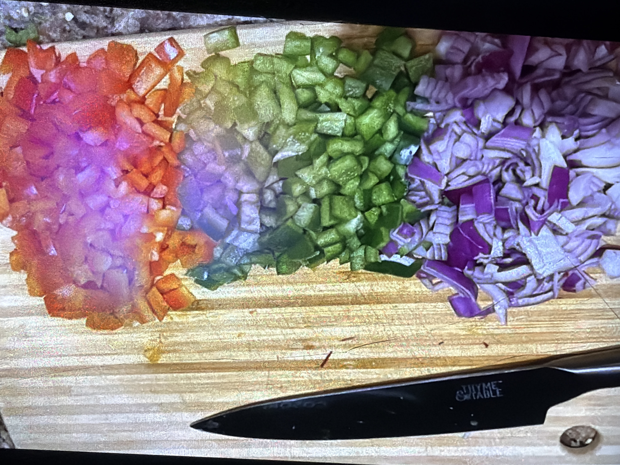The image shows a top-down view of chopped vegetables on a wooden cutting board. The vegetables are arranged in distinct piles, creating a gradient of colors. Starting from the top, there is a pile of chopped red bell peppers, transitioning into chopped orange bell peppers, then chopped green bell peppers, and finally a pile of chopped red onions at the bottom. A black knife with a silver handle is positioned on the left side of the cutting board, with its blade pointed downwards. The cutting board appears to be made of light-colored wood with visible grain patterns. The background is dark and out of focus, suggesting an indoor setting, possibly a kitchen. The lighting is somewhat uneven, casting subtle shadows and highlighting the textures of the chopped vegetables. There are no people visible in the image, and no specific details about the city of Bukuru, Nigeria, can be inferred from the scene.