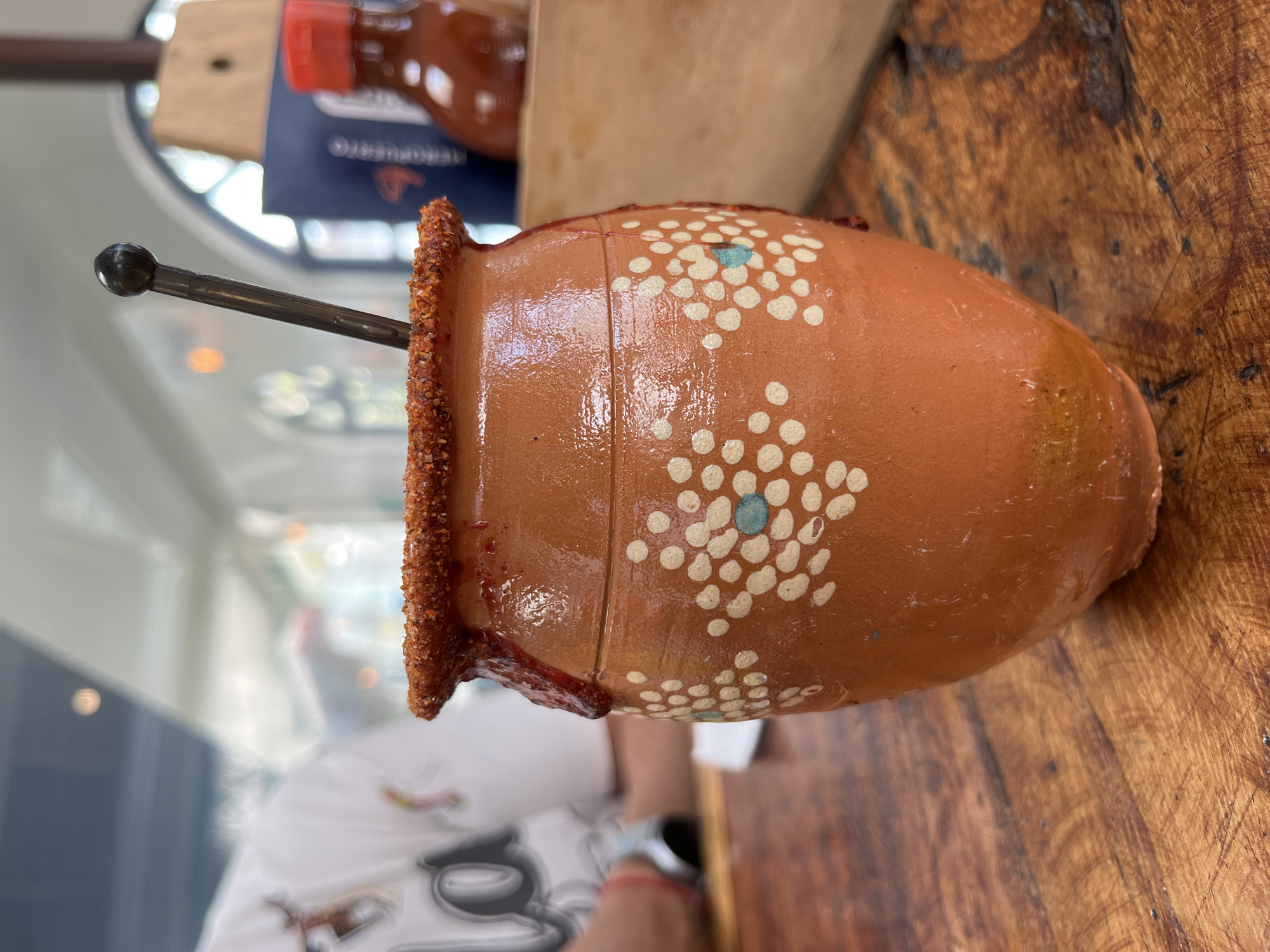 The image shows a clay cup with a chili-rimmed edge and a metal straw, decorated with white polka dots and a single turquoise dot. It sits on a weathered wooden table. Part of a white t-shirt and a person's arm with a watch are visible in the background. Behind the cup are blurred objects, including a bottle with a red cap and a menu, suggesting the location is a restaurant or bar. The city is Unknown, Unknown.