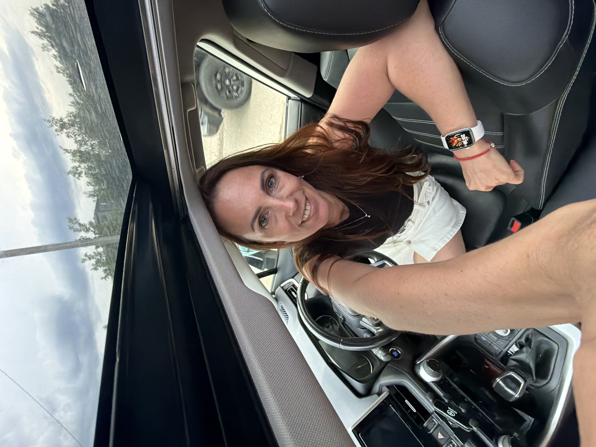 The image shows a woman sitting in the driver's seat of a car with a sunroof. She has long brown hair and is smiling at the camera. She is wearing a black top and white shorts. She has a smartwatch on her left wrist. The interior of the car is visible, including the steering wheel and dashboard. Outside the car, trees and a cloudy sky are visible. The location is not specified in the image, so I cannot confirm if it is in the city of Birobidzhan, Russia.