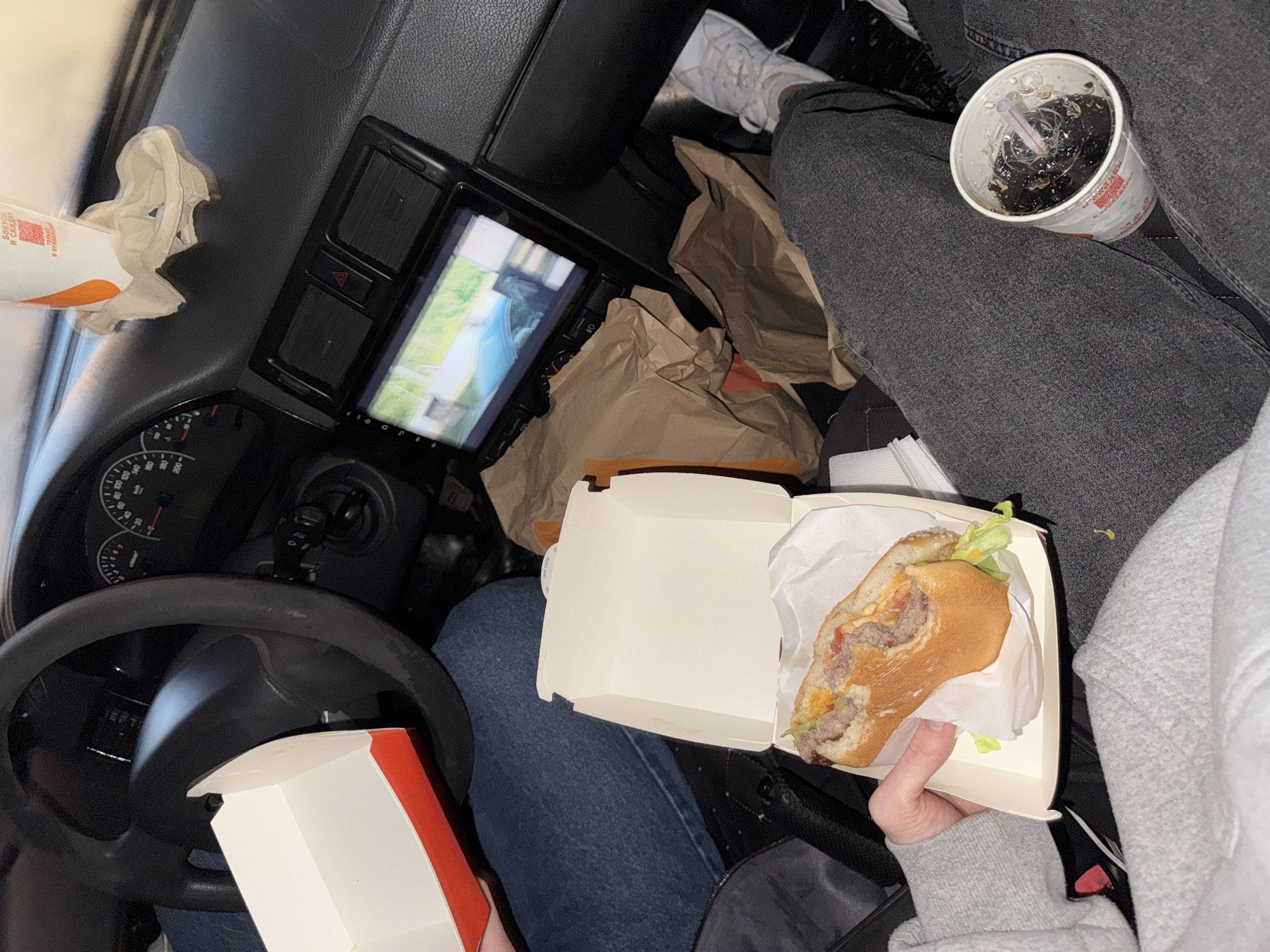 The image captures a candid, low-angle shot from inside a car, likely in Perm, Russia. The foreground is dominated by a person's legs, clad in dark grey jeans and a grey hoodie, resting on the car's floor and seat. To the right, a white sneaker is visible, suggesting another person is present. A white paper cup with a straw, filled with a dark liquid and ice, sits on the person's lap.
In the middle ground, a hand, likely belonging to the person in the foreground, holds an open burger box containing a half-eaten burger. The burger appears to have two patties, cheese, lettuce, and tomato. Another partially visible burger box is to the left. Behind the burger, several crumpled brown paper bags, consistent with fast-food packaging, are scattered across the car's interior.
The background reveals the car's dashboard, including the steering wheel, speedometer, and an infotainment screen displaying a video. A car's hazard light button is visible above the screen. A partially obscured fast-food drink cup, with a paper napkin underneath, is placed on the dashboard. The overall impression is one of a casual, possibly late-night meal being consumed inside a vehicle, with elements suggesting a fast-food experience in an urban environment.