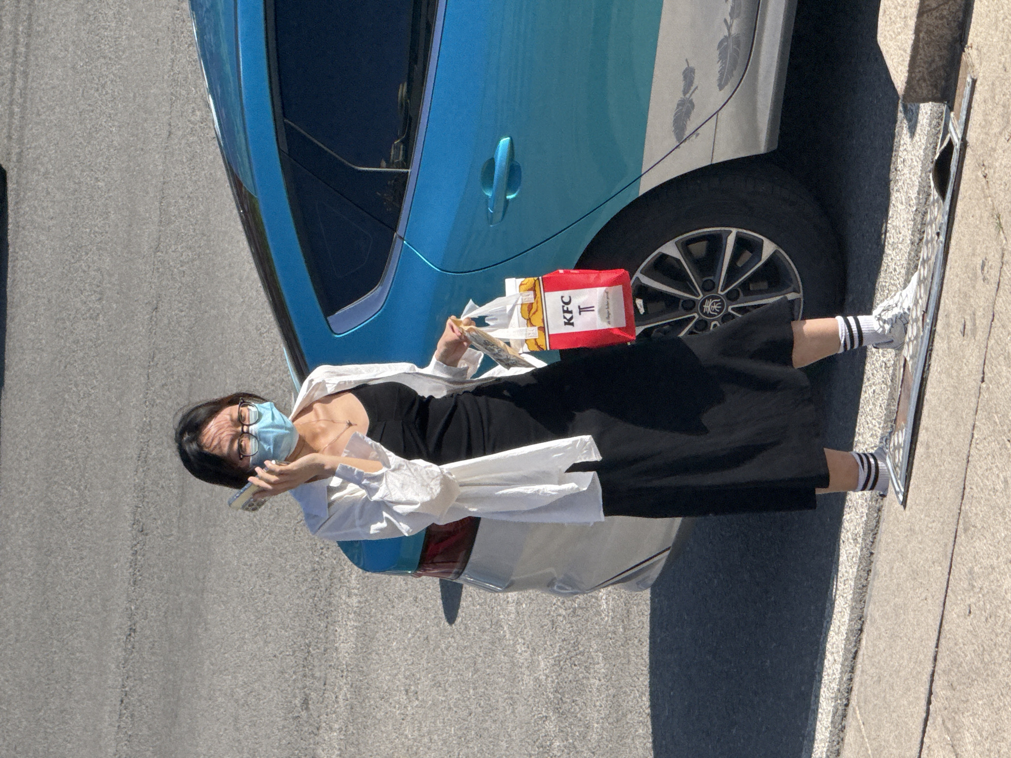 A person wearing a light blue disposable face mask, glasses, a black dress, white sneakers with black stripes on the socks, and a white shirt over the dress is standing on a sidewalk next to a light blue car. The person is holding a KFC bag and what appears to be a mobile phone. The car is parked next to the sidewalk. The setting appears to be a city street on a sunny day. The location is not specified.
