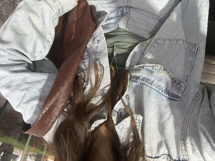 The image provides a close-up, chest-level view of an individual in an urban outdoor setting. The person is wearing a light-wash denim jacket, unbuttoned, over an olive green ribbed top and light-wash denim pants. A brown, textured shoulder bag, appearing to be crocodile or alligator embossed, is visible, draped across the person's left side. A silver necklace with a distinctive elongated, pointed pendant is centered on the individual's chest. The background, heavily blurred, indicates an environment consistent with a street or commercial area in Mexico City, Mexico, featuring indistinct reflections from glass storefronts or windows in the upper right and a dense, dark green hedge or landscaping element in the lower right. Blurry shapes in the far distance suggest urban structures or vehicles.