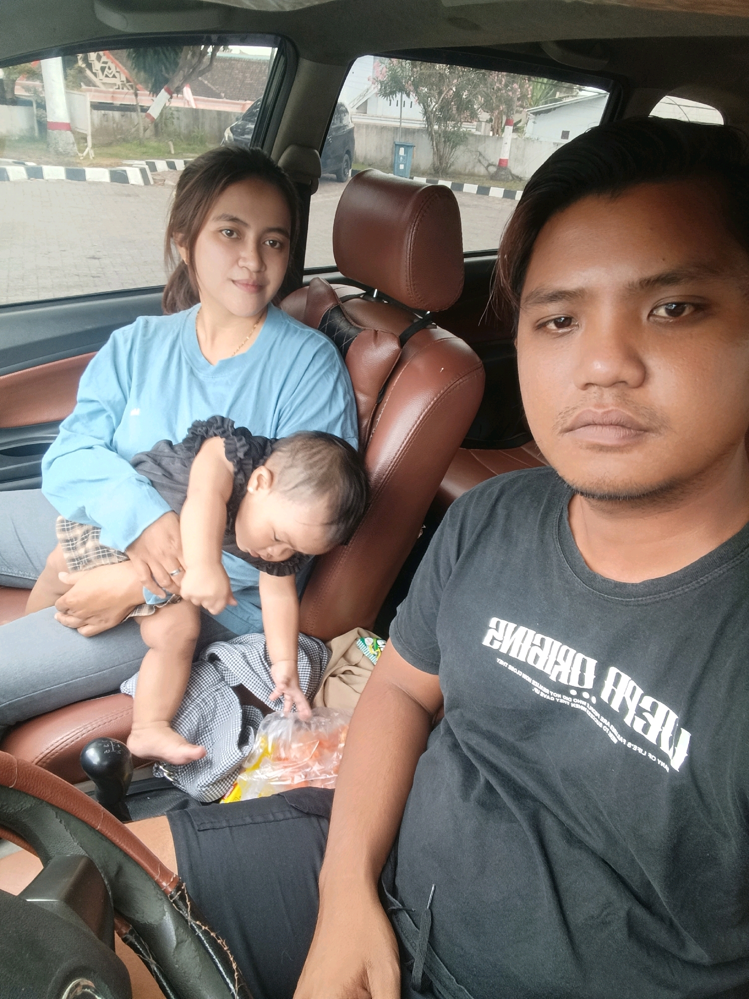 In Tempeh Lor, Indonesia, a man and a woman are seated inside a vehicle. The man is in the driver's seat, facing forward, while the woman is in the passenger seat, holding a baby. The baby is asleep on the woman's lap, dressed in a dark outfit with ruffled sleeves and a plaid skirt. The man wears a black t-shirt with white lettering, and the woman is in a light blue long-sleeved shirt and gray pants. The interior of the car shows brown leather seats and a gear shift. Outside the car, a paved area with a white and red striped barrier and some vegetation is visible.