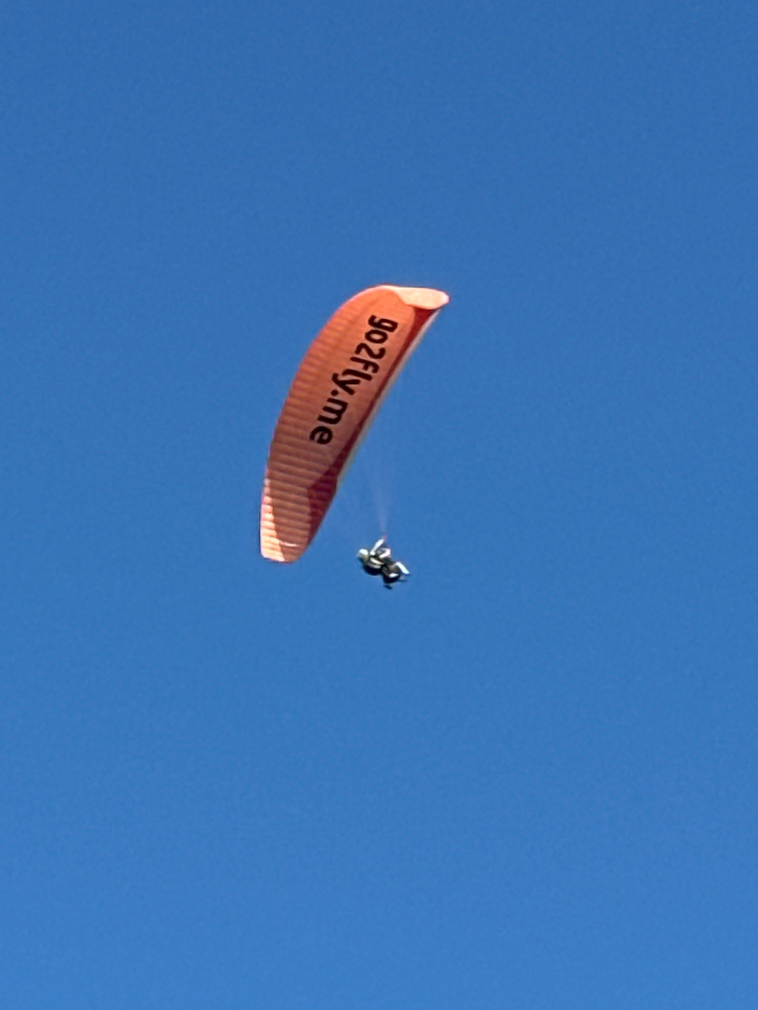 The image shows a paraglider in mid-air against a clear blue sky. The paraglider is orange and has the text "go2fly.me" printed on it. A person is suspended beneath the paraglider. The image appears to be taken from below, looking up at the paraglider. There is no indication of the city of Boreti, Montenegro, in the image itself.