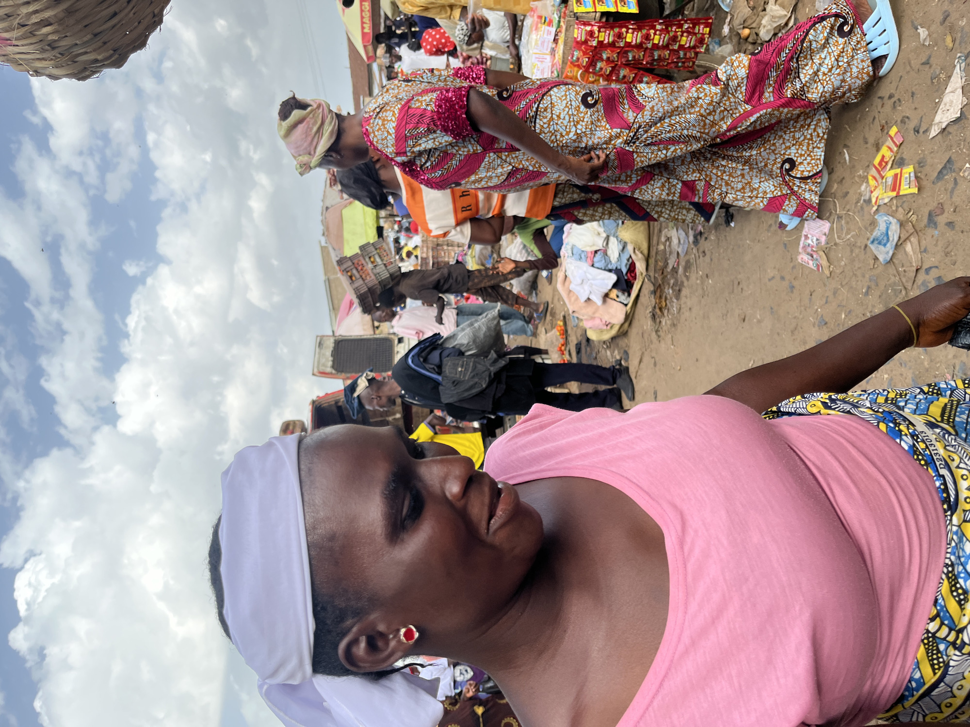 In Rafin Par, Nigeria, a busy marketplace scene unfolds under a cloudy sky. The foreground features a woman with a white headscarf and a pink t-shirt, adorned with red earrings, looking down and to her left, her face partially obscured. To her right, another woman stands in a vibrant, patterned dress and headscarf, her gaze directed away from the camera.
The marketplace is a bustling environment filled with people and goods. In the background, individuals are seen walking and interacting, some carrying loads. Crates of produce are stacked, and market stalls are visible, suggesting a lively commercial activity. The ground is littered with debris and discarded items, typical of a crowded outdoor market. The overall impression is one of everyday life and commerce in a Nigerian city.
