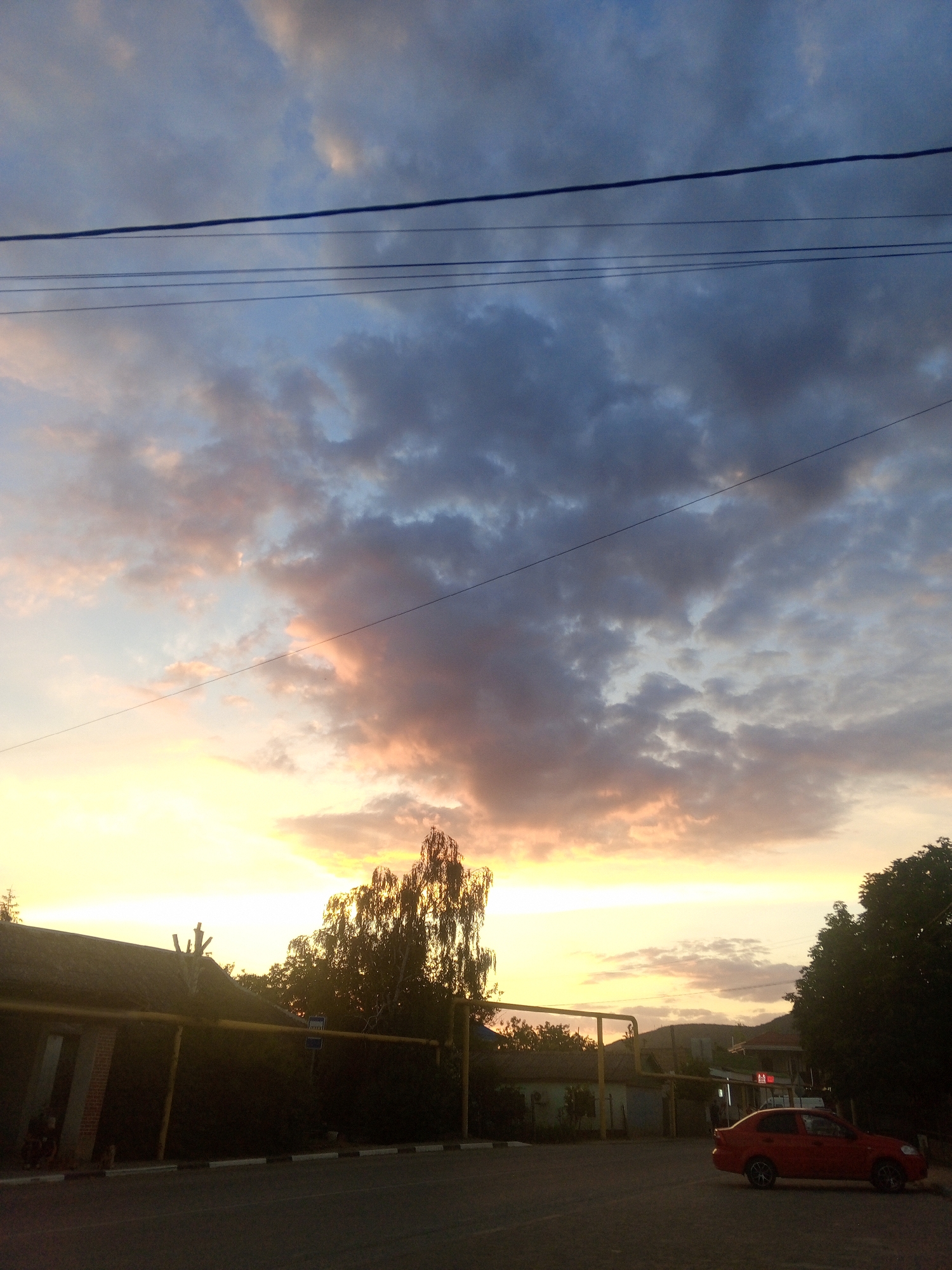 The image captures a sunset over the village of Golubinskoye, Ukraine. The sky is filled with dramatic clouds, ranging from dark grays to soft pinks and oranges, illuminated by the setting sun. A prominent line of power lines stretches across the upper portion of the frame, with thinner wires below. In the foreground, a paved road is visible, with a red car parked on the right side. To the left, the roofline of a building, possibly a house or a small commercial establishment, is partially obscured by trees. The trees themselves are silhouetted against the bright sky. The overall scene depicts a rural or semi-rural setting at dusk.