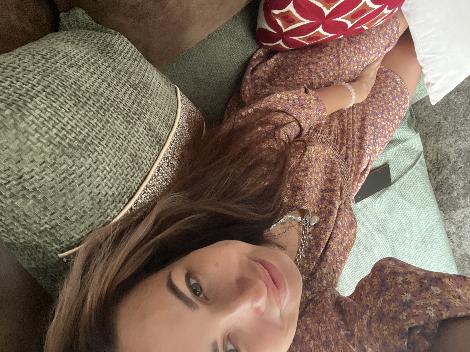 A woman in a floral dress is taking a selfie on a couch. She has brown hair and is wearing a necklace and a bracelet. The couch is green and has several pillows. A phone is visible on the couch.