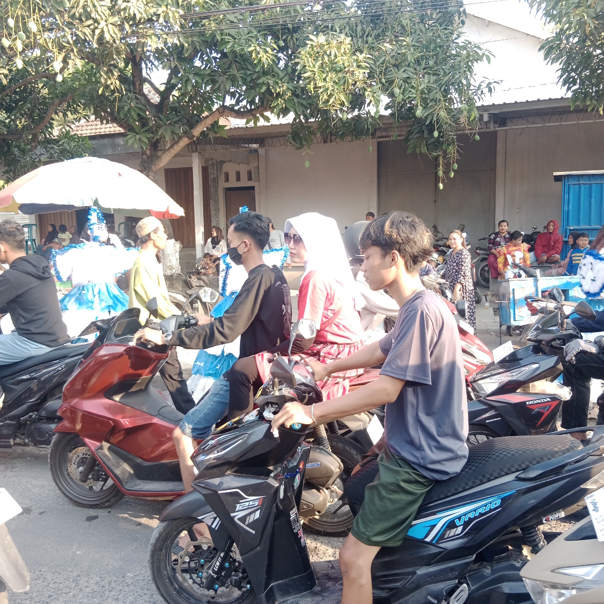 In the city of Tegalrejo, Indonesia, an outdoor scene depicts a street with several people and motorbikes. A large tree with ripening mangoes shades the area. In the foreground, two young men are seated on motorbikes. The man on the left is wearing a black hoodie and blue jeans, his face obscured by a black face mask. He is looking forward and appears to be waiting. The man on the right, wearing a grey t-shirt and green shorts, is also seated on a motorbike, with his right hand resting on the handlebars. His gaze is directed to his right.
Behind them, a woman in a white hijab and a red and white checkered skirt is also on a motorbike. She wears sunglasses and looks to her left. To her left, a person in a black shirt and blue jeans is seated on a motorcycle. Further back, a person in a blue and white elaborate costume, possibly part of a celebration, stands under a colorful umbrella. Several other individuals are visible in the background, some seated on motorbikes and others standing, suggesting a gathering or event. Buildings with white walls and corrugated roofs line the street. The overall impression is a bustling street scene with a mix of everyday life and celebratory elements.