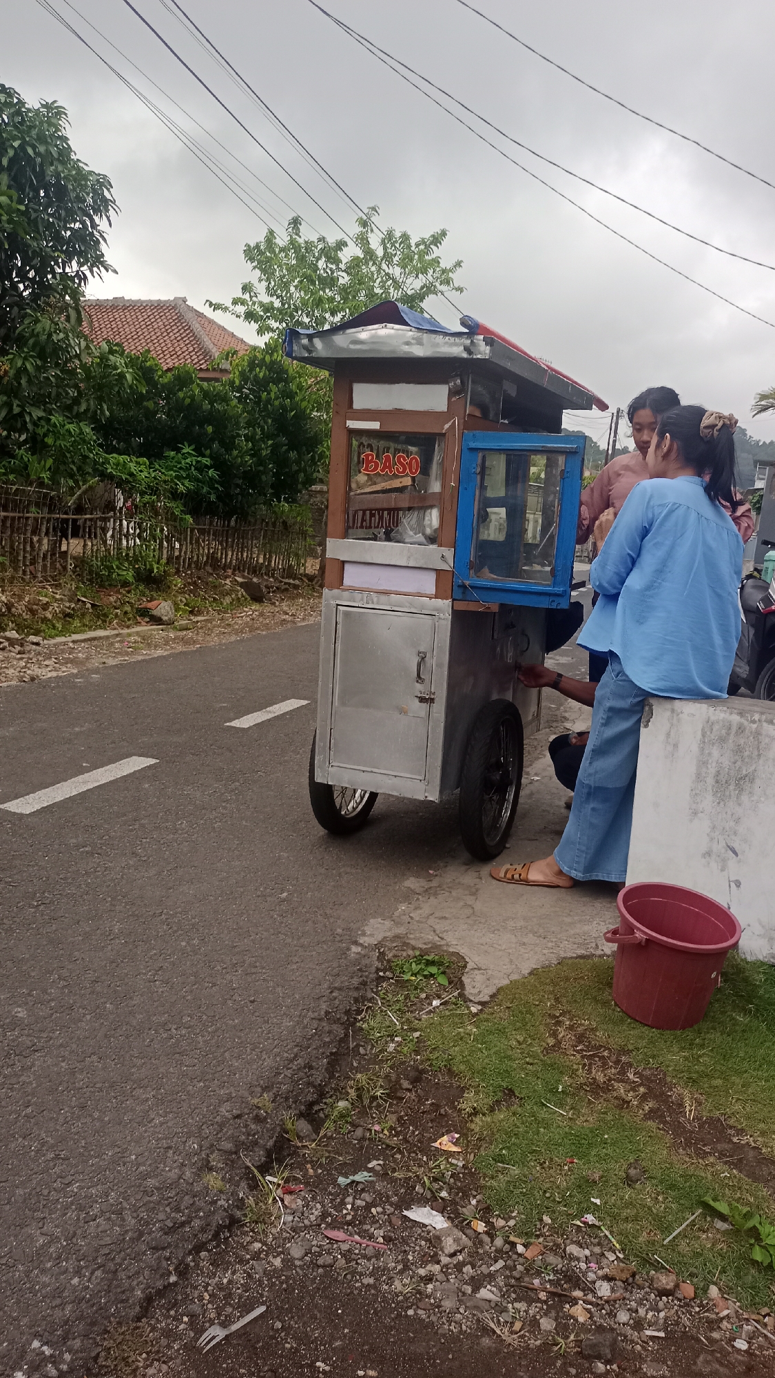 In Pakemitan, Indonesia, a scene unfolds on the side of a road with a grey, overcast sky. In the foreground, a paved road with a white dashed line runs from left to right. To the right of the road, there's a patch of grass littered with small stones, debris, and a pink plastic spoon. A maroon plastic bucket sits on the grass. A concrete barrier runs along the edge of the grass.
Dominating the center is a food cart, designed for selling "BASO" (meatballs). The cart is constructed with a wooden frame and metal siding, mounted on two spoked wheels. It features a glass display case with blue framing, and a metal door on the lower section. Inside the glass case, some items are visible, and the word "BASO" is clearly displayed on the wooden part above.
Three individuals are interacting around the food cart. One person, wearing a light blue long-sleeved shirt and blue pants, is seated on the concrete barrier, facing the cart. Another person, in a pink top, stands near the cart, and a third person's hands are visible reaching into the cart from behind.
The background reveals a residential area with a house featuring a red-tiled roof, surrounded by lush green trees. Overhead, multiple power lines are strung across the sky. The overall impression is a casual street-side food vendor scene in a semi-urban or rural Indonesian setting.