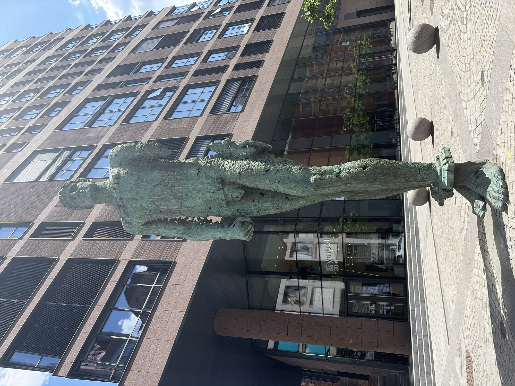 The image shows a full-body bronze statue of a male figure standing in front of a modern, multi-story building with a reddish-brown facade and numerous glass windows. The statue is greenish-bronze in color and appears to be weathered. It is positioned in the foreground, dominating the lower half of the frame. The building rises behind the statue, its facade a grid of windows and darker vertical elements. A large glass-fronted shop is visible at the base of the building, with the word "symbol" and "30+" legible on the window. Several large, spherical stones are placed on the paved ground in front of the building and around the statue. The sky is partly cloudy, with bright sunlight casting shadows. The scene appears to be an urban outdoor space. There are no people visible in the image.