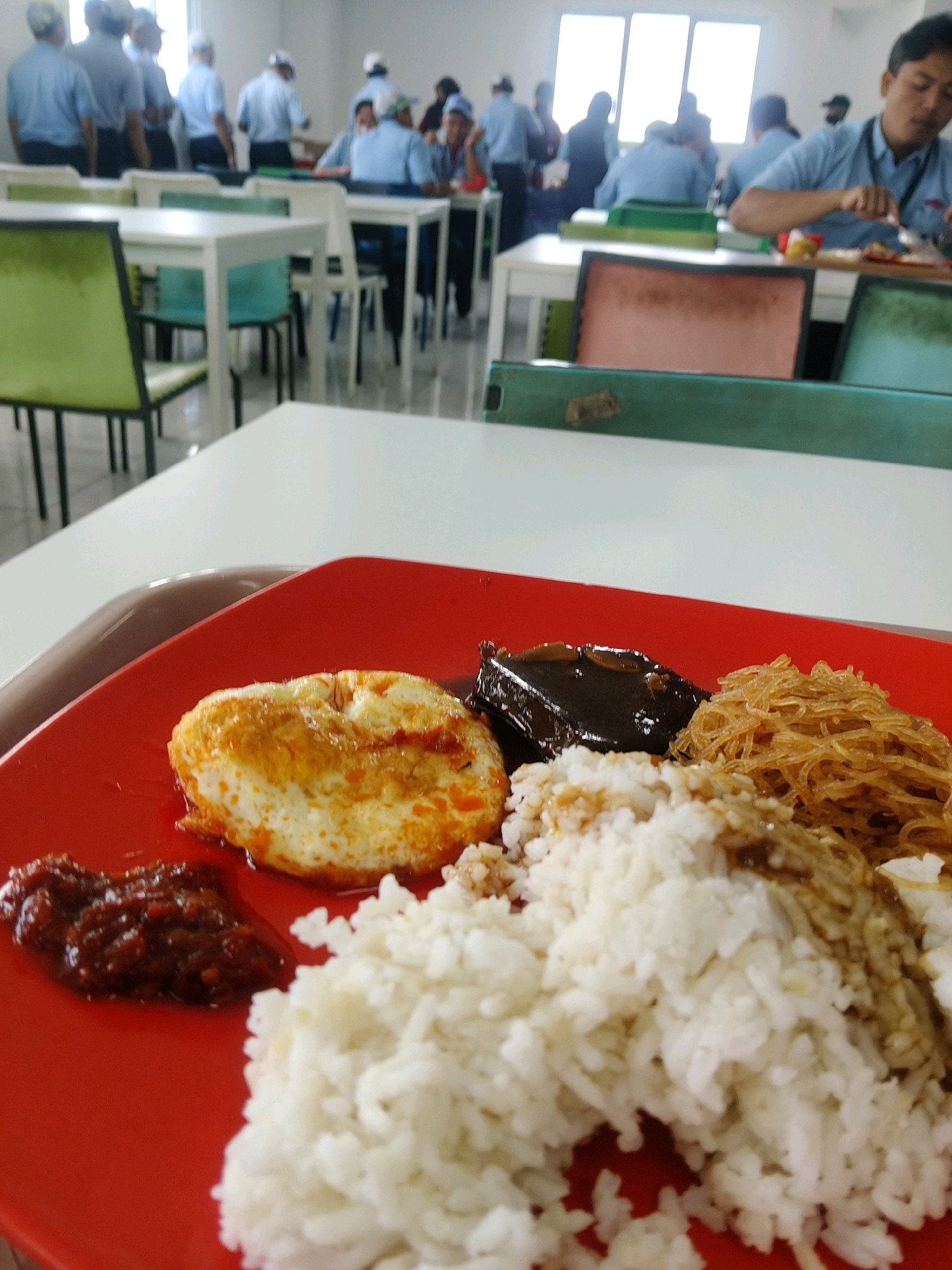 The image captures a close-up of a red plate of food, likely in a canteen or cafeteria setting in Mekarmukti, Indonesia. The food consists of a portion of white rice, a fried egg with a runny yolk, a dark, saucy item, and a pile of shredded, golden-brown noodles. To the left of the rice, a small dollop of red chili paste is visible.
In the blurred background, several people wearing light blue uniforms and white caps are seen standing and sitting at tables. The environment appears to be an indoor space with large windows, suggesting a common area or dining hall. Some tables are white with green chairs, while others are partially obscured. The overall impression is a typical scene of a mealtime in a workplace or institutional setting.