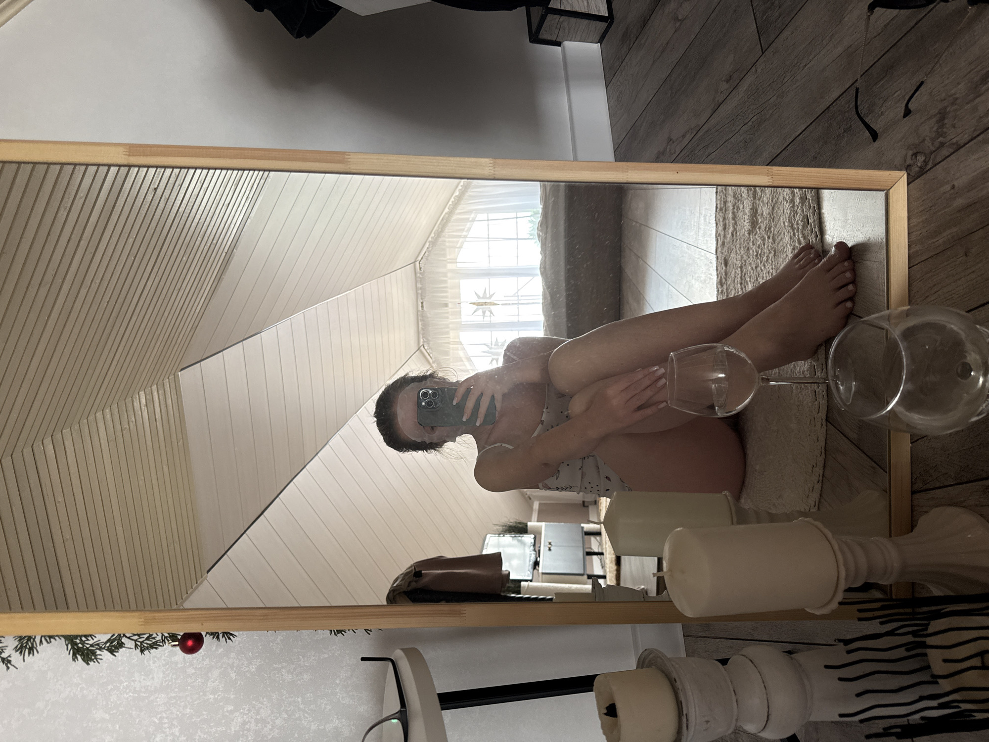 The image shows a person reflected in a rectangular mirror with a light brown wooden frame. The mirror is positioned indoors, likely in a residential setting. The person in the reflection is holding a mobile phone, partially obscuring their face. They are sitting with their legs crossed. Two wine glasses are visible near the lower right corner of the mirror. A white candle and decorative objects are visible as well. The mirror reflects a room with white paneled walls and a window with a decorative star pattern. The floor appears to be made of wood planks. The presence of a Christmas ornament at the edge of the mirror implies that the photo was taken during the holiday season. The exact location is not evident, but the prompt mentions the city of Chaikovsky, Russia, suggesting that this is the location.