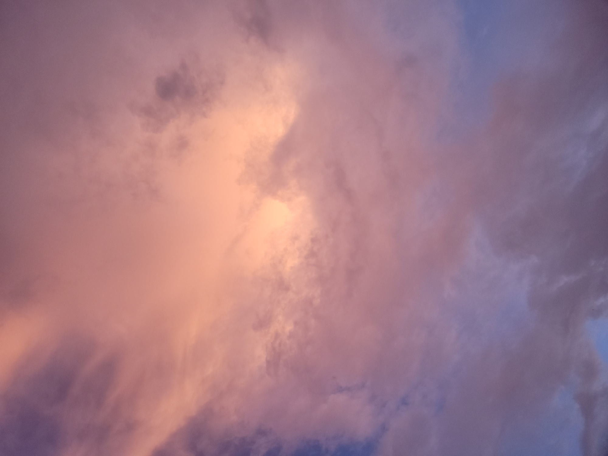 The image presents a vibrant and dramatic cloudy sky, likely captured during either sunrise or sunset. The sky is dominated by a rich palette of warm colors, primarily peach, orange, and pink, which transition smoothly into cooler shades of purple and blue towards the edges of the frame.
A soft, golden light illuminates the clouds from within, casting a warm glow that highlights their contours and textures. Some cloud formations appear dense and voluminous, showcasing darker, shadowed undersides contrasting with brightly lit peaks. Other areas feature thinner, more diffused cloud cover, allowing hints of the deeper blue sky to emerge. The overall effect is one of an ethereal and captivating atmospheric display.
There are no discernible people, objects, or visible text within the frame. The scene depicts a natural phenomenon, emphasizing the beauty and changing colors of the sky during a twilight hour. Given the context, this tranquil and colorful sky is observed over Jos, Nigeria.