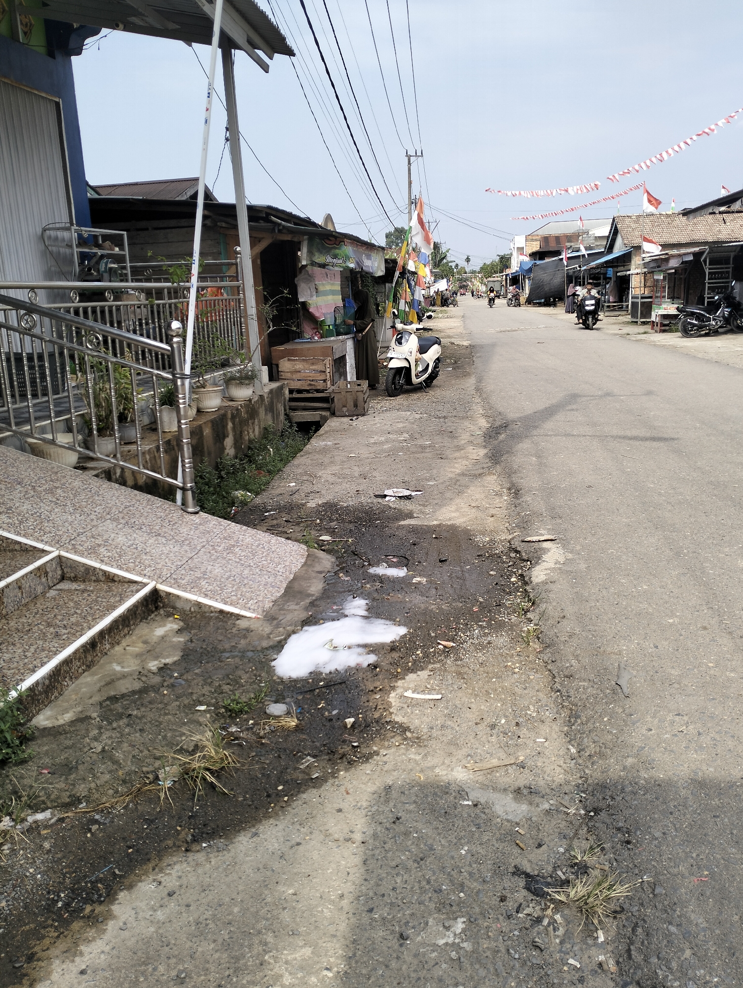 The image depicts a street scene in Pematang Lumut, Indonesia. The foreground shows a concrete sidewalk with scattered debris, including a patch of white foamy substance. A ramp with a tiled surface leads up to a building on the left. The street itself is paved asphalt, with a dirty shoulder. Several motorcycles are parked on the side of the road, and others are being ridden by people. Buildings with corrugated metal roofs line the street, some appearing to be shops. Red and white Indonesian flags and pennants are visible, suggesting a national celebration or event. The sky is overcast.
