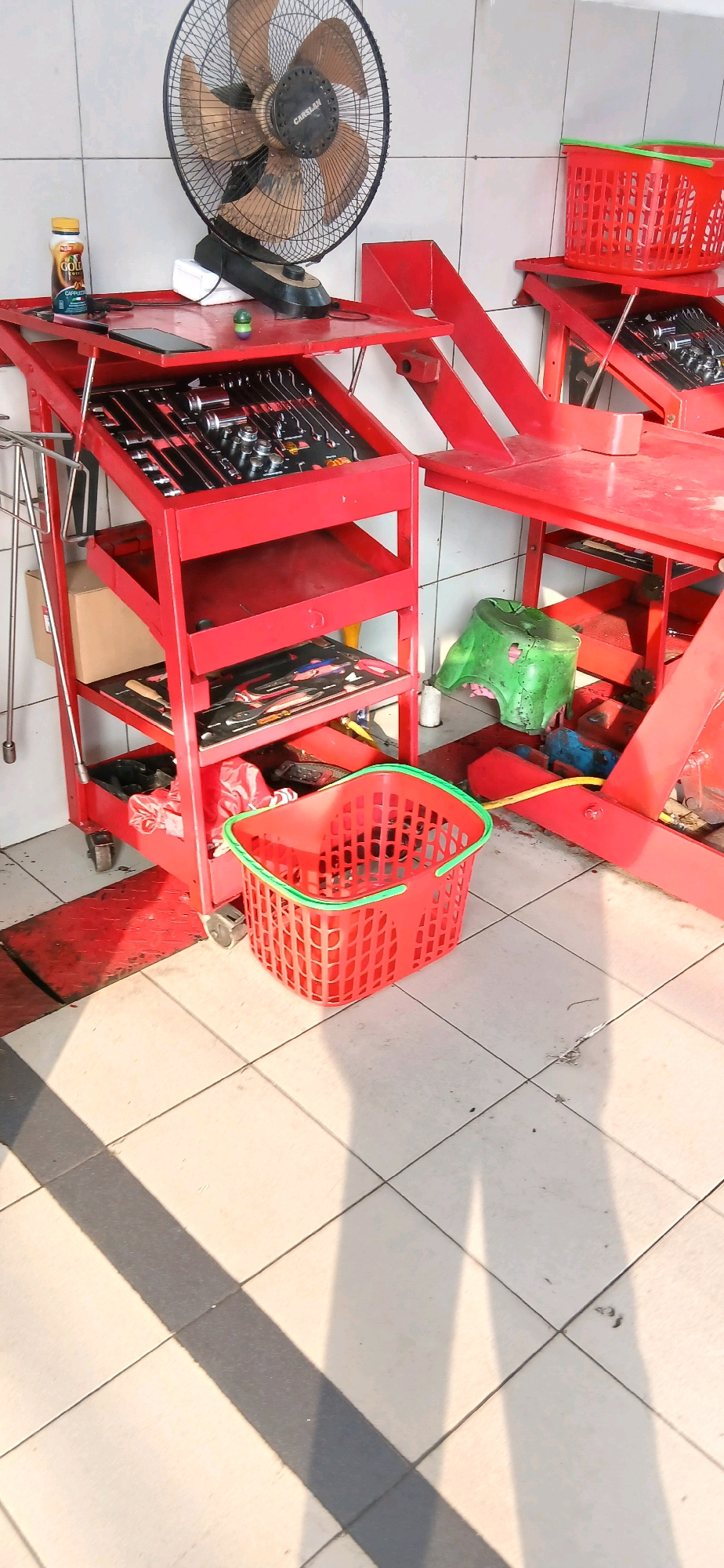 This image shows a red tool cart with various tools, a fan, and a bottle on a shelf. On the floor, there is a red basket with a green rim. The scene appears to be inside a workshop or garage. The tiled floor and the lighting suggest an indoor environment, possibly in Samarinda, Indonesia. The image is taken from a slightly elevated angle, looking down on the tool cart and surrounding area.