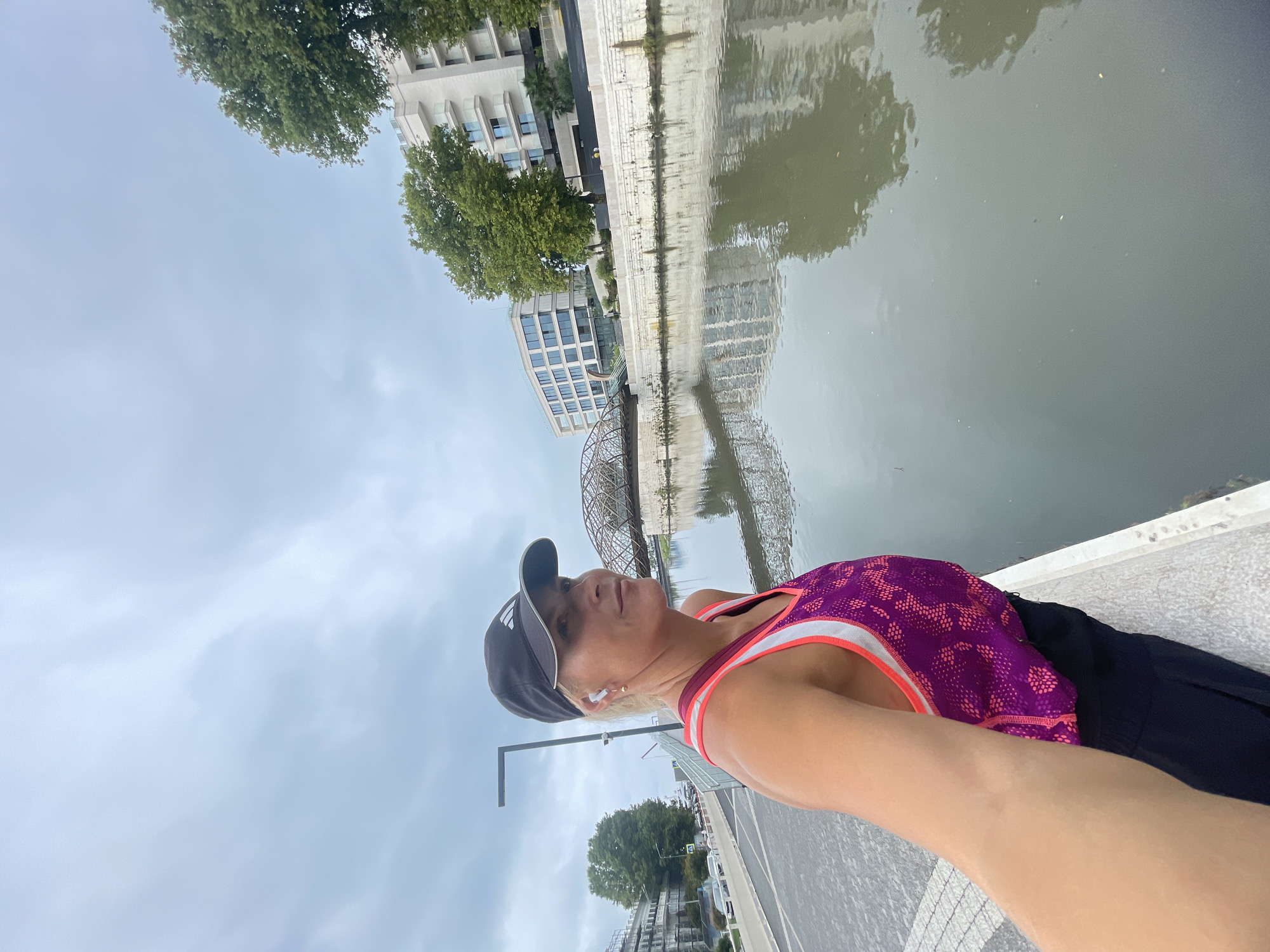 A blonde woman wearing a black baseball cap and a purple and pink athletic top is standing on a paved walkway next to a calm body of water. She is looking to her right. Her right arm is extended, holding the camera. In the background, there are modern buildings, trees, a bridge, and a cloudy sky. The scene appears to be in an urban environment along a waterfront. The location is the waterfront in Sochi, Russia.