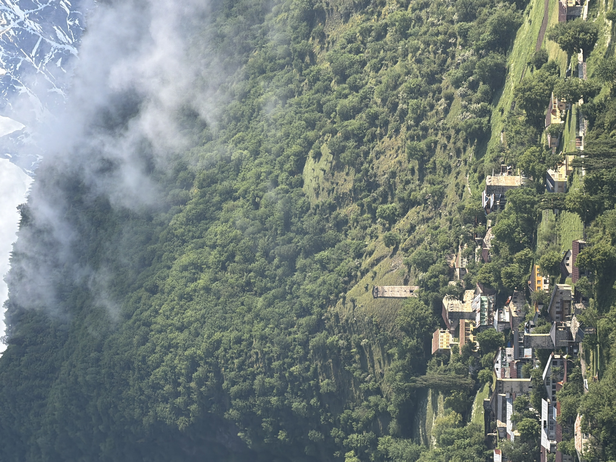 A vertical, high-angle shot depicts the mountainous landscape of Mestia, Georgia. The scene is dominated by a lush green mountain slope covered in dense trees. Patches of white fog drift across the upper portion of the mountain, partially obscuring the snow-capped peaks visible in the far distance. Nestled on the mountainside is a small village. The architecture of the village features stone houses and distinctive Svan towers, which are tall, slender defensive structures. The towers are a prominent feature of the landscape, appearing scattered throughout the settlement. The village is surrounded by trees and grassy slopes, with a road visible in the lower portion of the image. The overall impression is one of a remote and historic settlement within a dramatic natural setting.