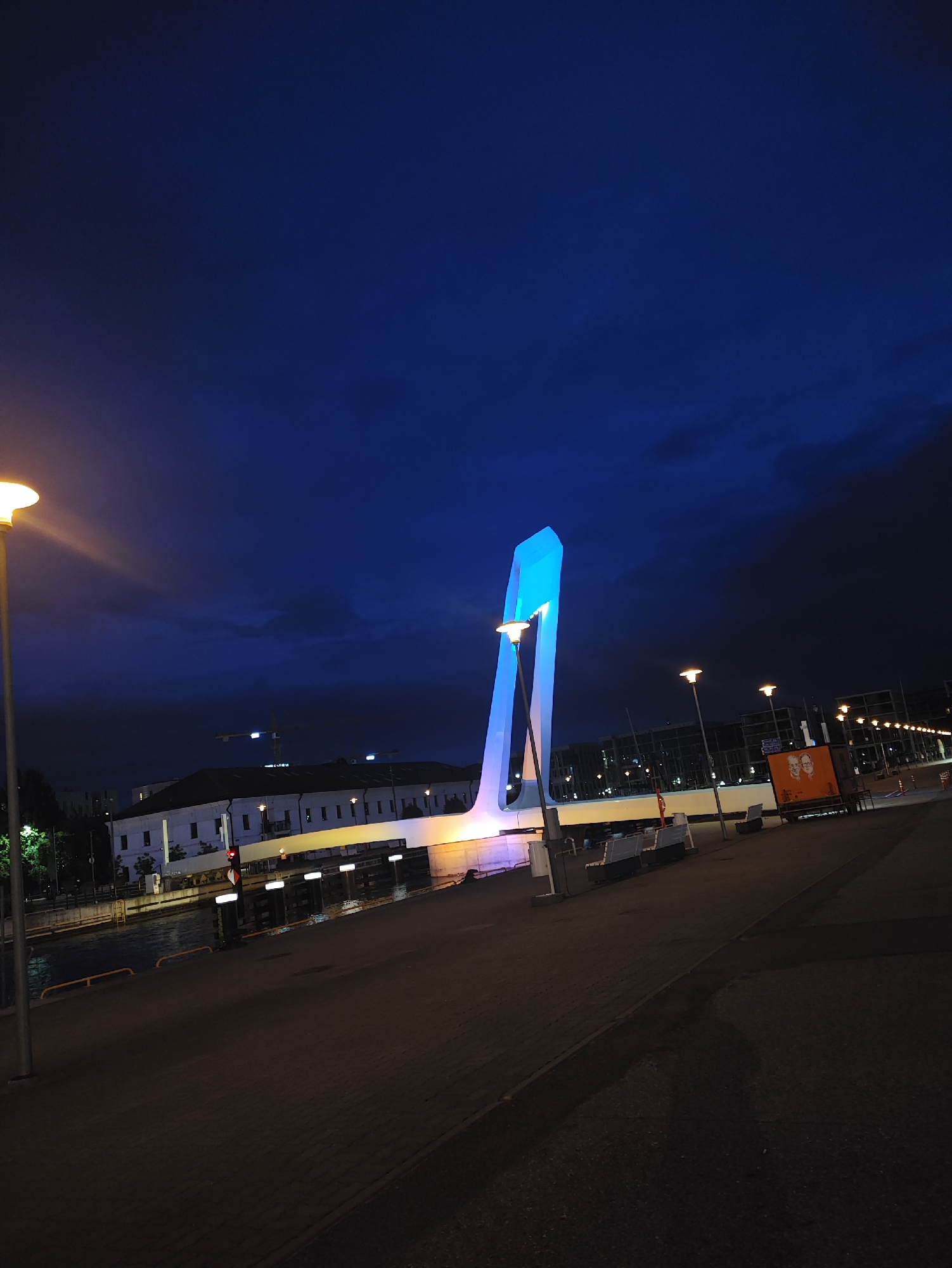 The image depicts a nighttime scene of a modern bridge illuminated with blue light in Tallinn, Estonia. The bridge features a distinctive, upward-sweeping design. To the left, a lamppost with a warm yellow glow stands on a paved walkway next to a body of water. Across the water, a white building with multiple windows is visible, with a construction crane in the background. In the foreground, the walkway is dark, with several streetlights spaced along its edge. To the right of the bridge, a bright orange sign with two portraits is mounted on a stand. The sky is a deep, dark blue with some cloudy textures.