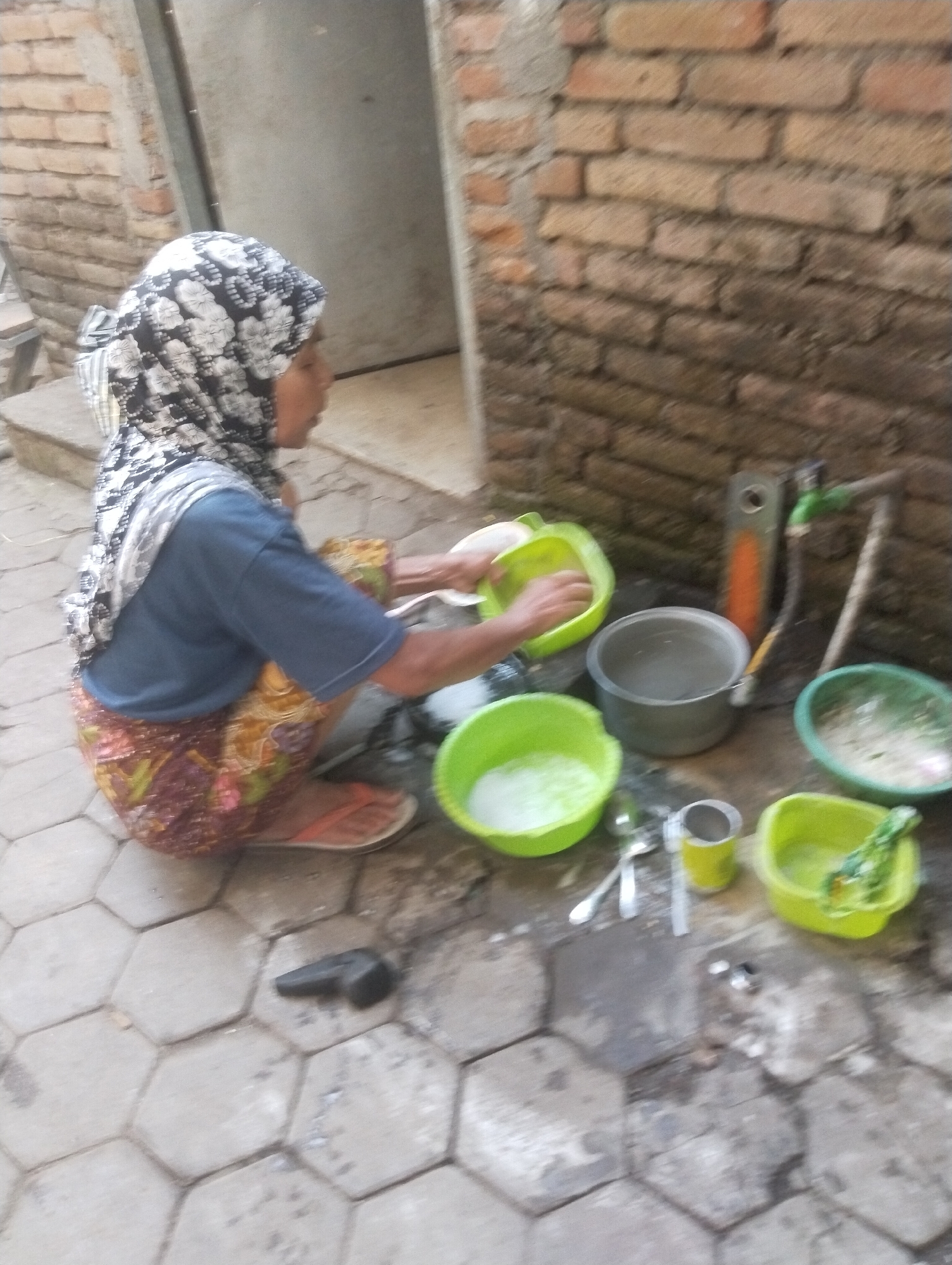In Wanasaba Daya, Indonesia, a woman is seen squatting on a hexagonal paved ground in front of a brick wall. She is wearing a blue shirt, a patterned skirt, and a headscarf. She is engaged in washing dishes in several green plastic basins. One basin contains soapy water, and she appears to be rinsing a dish. There is also a metal pot, a faucet attached to a pipe, and other miscellaneous items on the ground. The scene suggests an outdoor washing area.