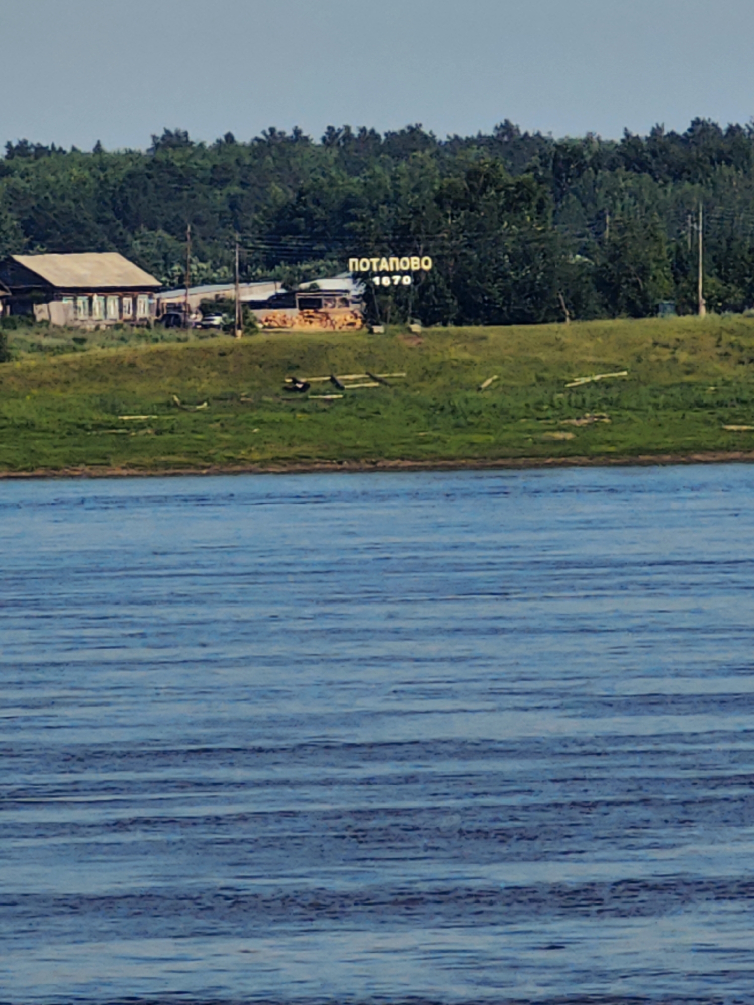 The image shows a wide, calm body of water in the foreground. In the midground, a grassy embankment slopes upwards towards a settlement. On the embankment, there is a cluster of buildings, including a traditional wooden house with a light-colored roof and several windows. To the right of the house, there are some smaller structures and what appear to be stacks of lumber or wood. Above the settlement, a sign with Cyrillic lettering that reads "ПОТАПОВО 1670" is visible, indicating the location is Potapovo, Russia, and that the settlement dates back to 1670. Behind the settlement, a dense line of green trees forms the background under a clear, pale blue sky. The overall scene depicts a rural settlement situated along a river.