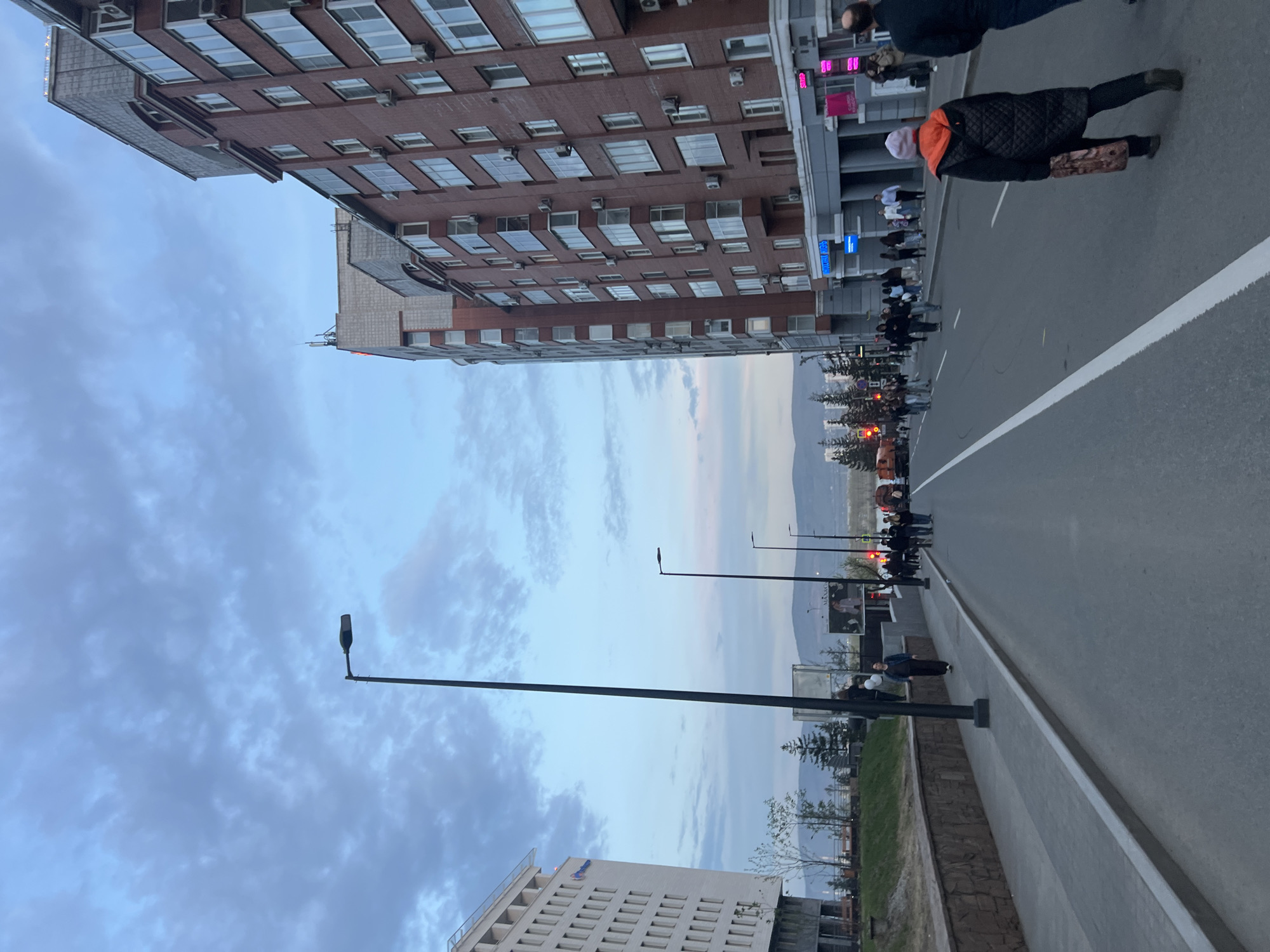 The image shows a street scene in Krasnoyarsk, Russia. A red brick building with numerous windows stands on the left. The street is wide with a white dividing line. Pedestrians are walking on the sidewalk. There are streetlights on the side of the road. In the background, there are trees and mountains. The sky is cloudy. Another building with white walls is in the lower left corner.