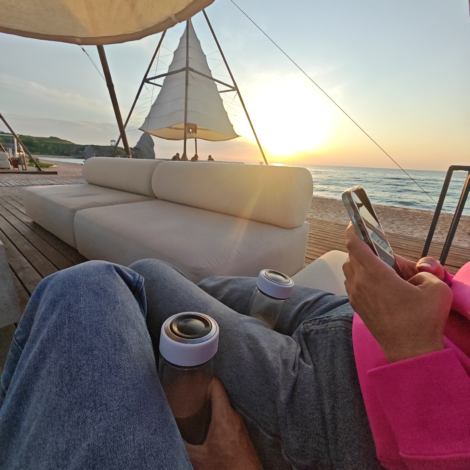 The photograph shows a first-person perspective of individuals relaxing on a beachfront lounge area in an unidentified location. The time appears to be sunset, with the sun low on the horizon over the sea. Two people are visible from the knees down. One person is wearing blue jeans, and the other is wearing grey jeans and a bright pink jacket. Two clear bottles with white lids are sitting on their laps. One of the people is holding a smartphone in their hands, seemingly using it. In the background, there is a white lounge sofa on a wooden deck, and a structure resembling a sail is visible, casting shadows. Beyond the deck, there is a beach and the sea. The lighting is warm due to the sunset.
