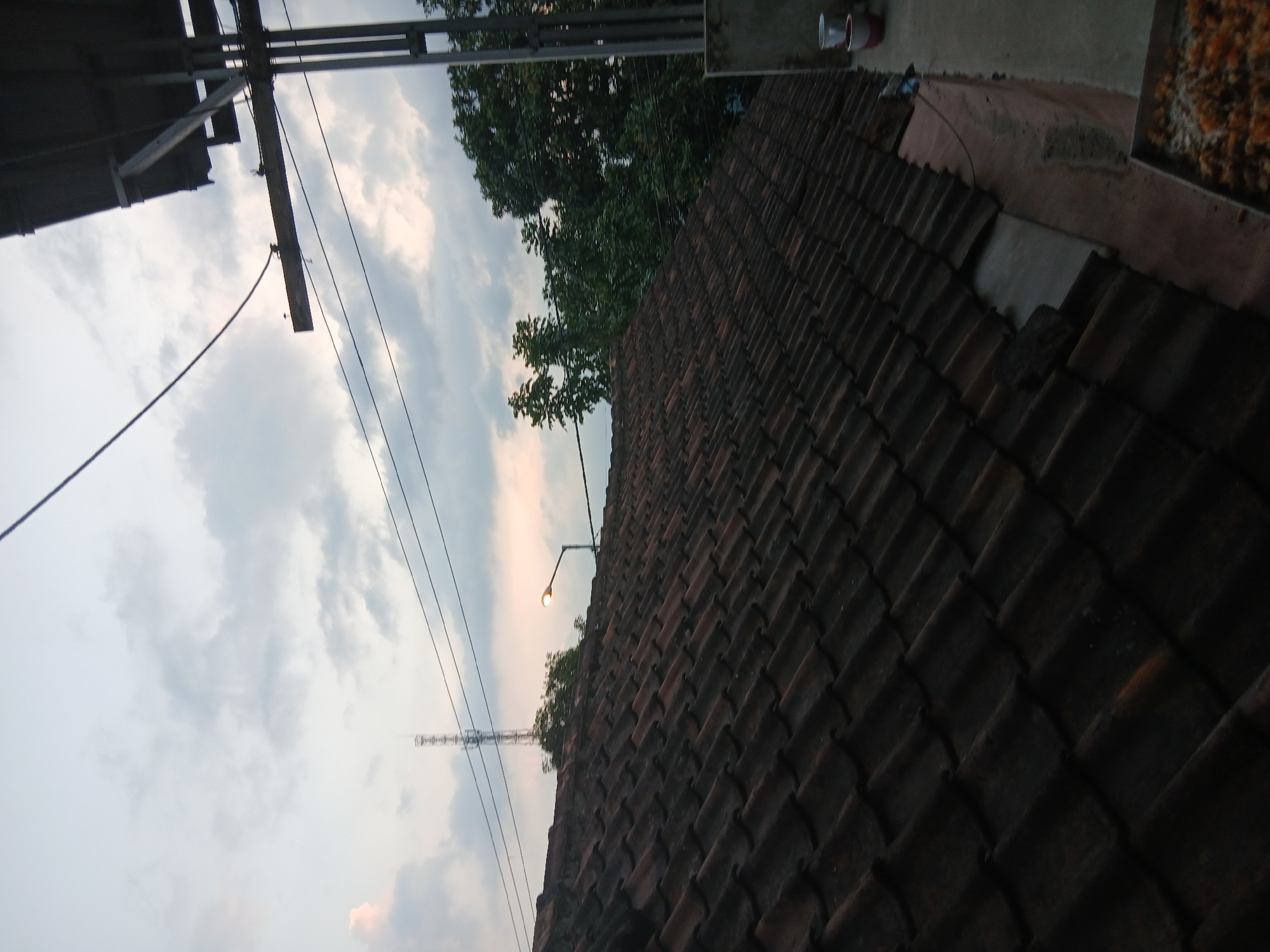The image depicts a view from a tiled roof, likely in Kademangan Kulon, Indonesia, during twilight. The sky is overcast with soft, diffused light, suggesting either dawn or dusk. In the background, a tall communication tower is visible, along with lush green trees. A street light is illuminated, casting a warm glow. The foreground is dominated by a dark, reddish-brown tiled roof, which appears somewhat aged. To the right, a concrete ledge is present, with two buckets visible, one white and one red and white. A metal tray filled with what appears to be dried food items or snacks is also present on the ledge. Electrical wires crisscross the scene, typical of an urban or semi-urban Indonesian environment. No people are visible in the image.