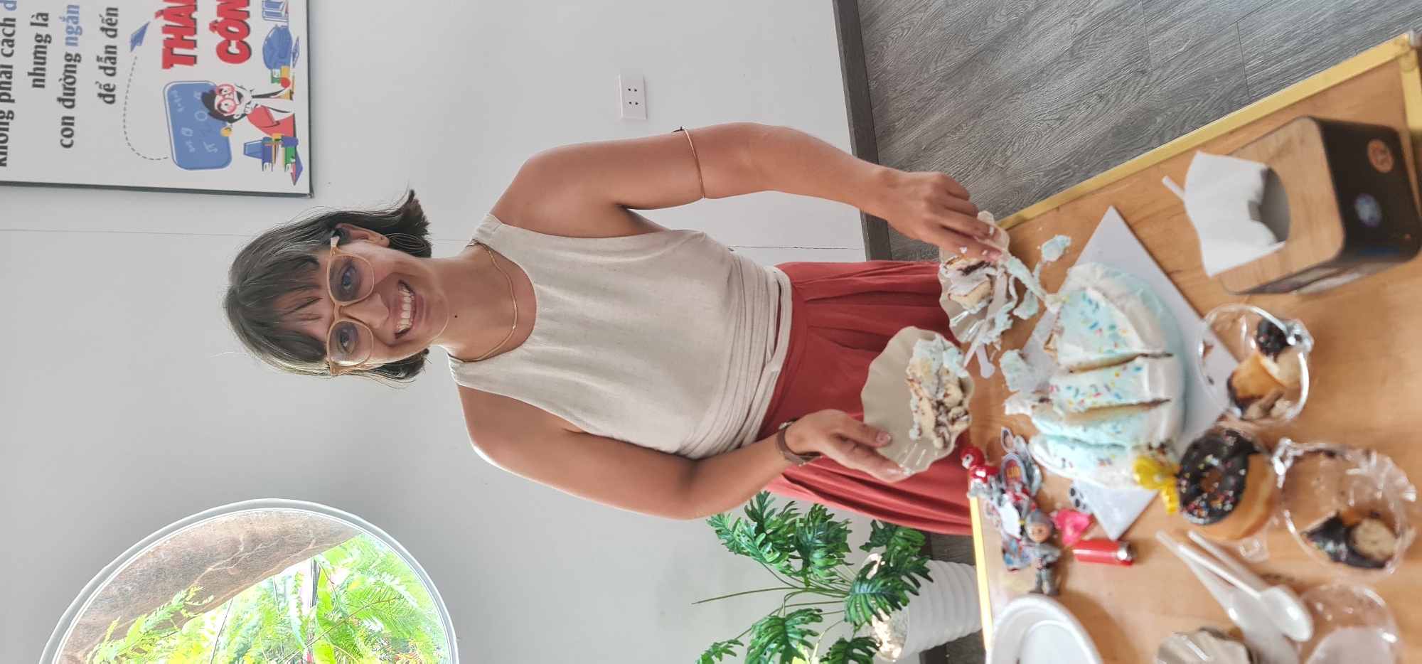A woman is standing in what appears to be an indoor setting, possibly an office or common area. She has short, dark hair, wears glasses, a light-colored sleeveless shirt, and red pants. She is holding a plate with a piece of cake on it, and there is another piece of cake on the table in front of her. The cake has white frosting with blue and multicolored sprinkles. The table also contains donuts, plastic containers with pastries, a tissue box, and some small toys or figurines. Behind the woman to the left is a large circular window showing green foliage outside, suggesting a garden or outdoor space. Behind her to the left is a sign with text in Vietnamese. The image also includes a plant in a white pot. The room has a gray floor and white walls. Overall, it appears to be a casual gathering with cake and other treats.