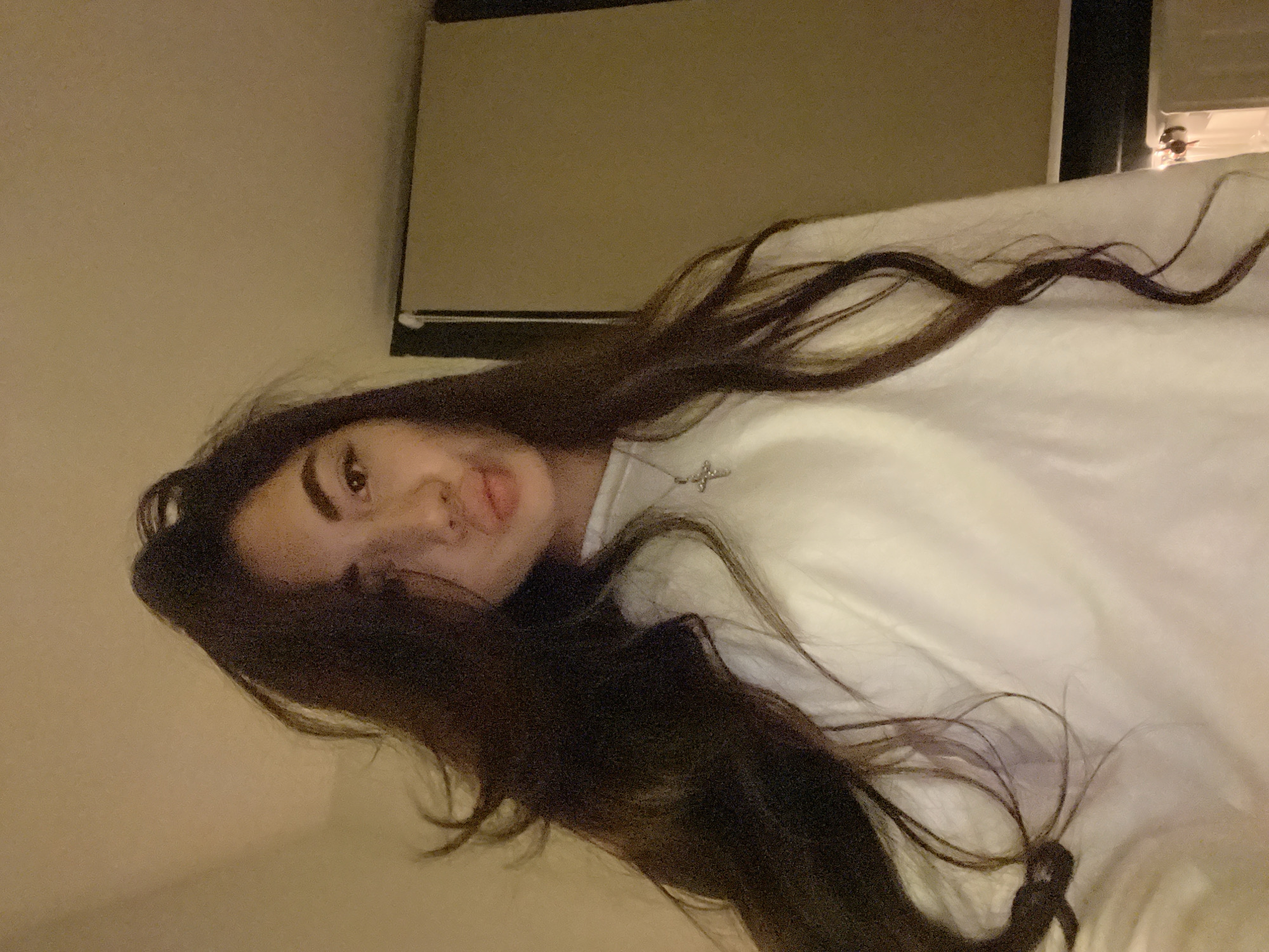 A young woman with dark, long, wavy hair is captured in a close-up, selfie-style photograph. She is wearing a white crew neck sweatshirt and a silver cross pendant necklace. Her lips are slightly pursed, and her gaze is directed towards the camera. The background is dimly lit and appears to be an indoor setting, possibly a room, with a textured wall and a partially visible projection screen or window covering. There is no discernible indication of the city of Chotěšov, Czechia.