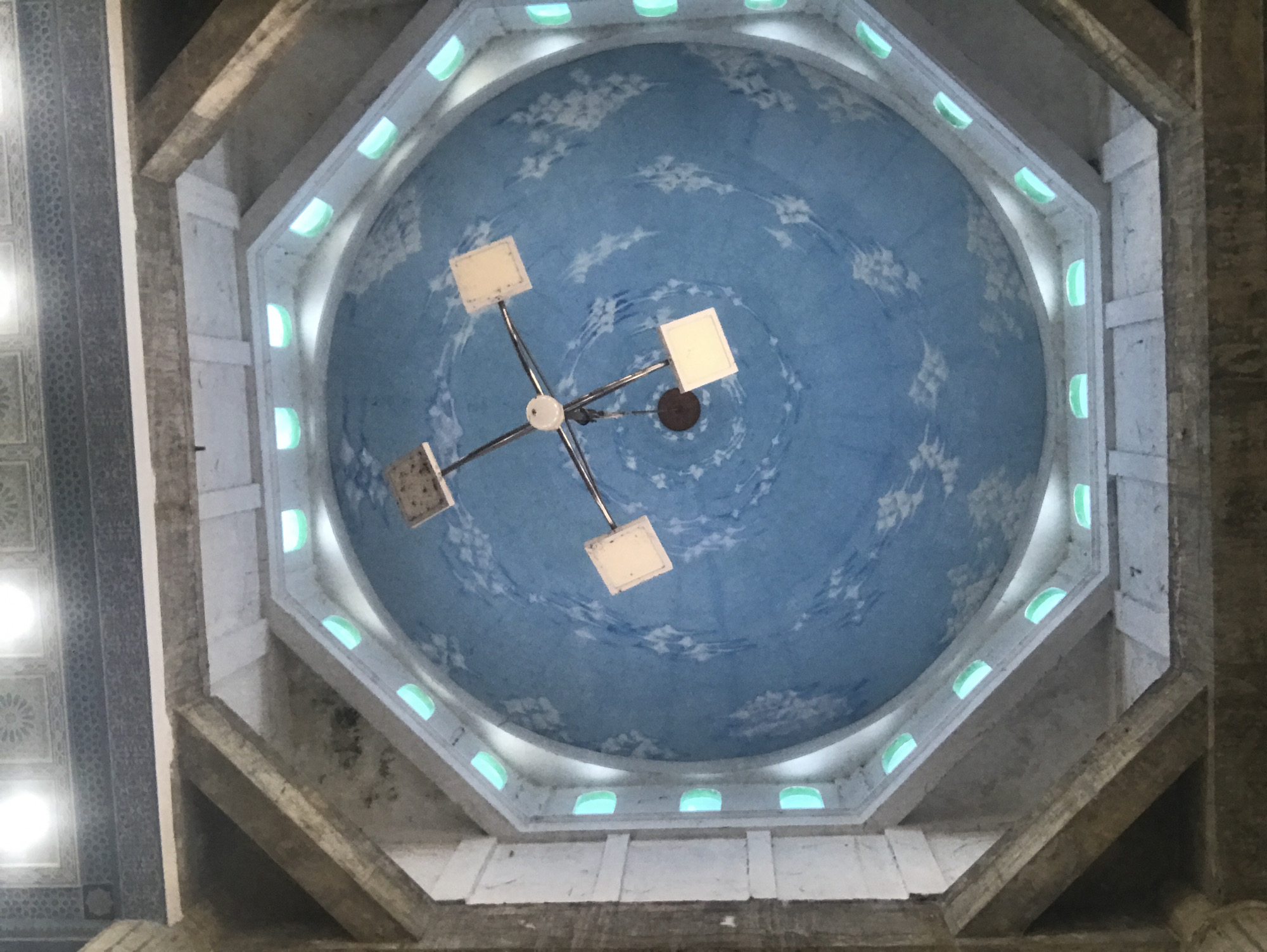 The image shows the interior of a building in Lhokseumawe, Indonesia, specifically looking upwards at a domed ceiling. The dome is painted a light blue with white cloud-like patterns. A modern chandelier with four square light fixtures hangs suspended from the center of the dome. The dome is surrounded by an octagonal structure with small, arched windows spaced around its perimeter. The surrounding structure is made of light-colored stone or concrete. Portions of the building's outer structure, also constructed of stone or concrete, are visible in the corners of the image. A section of decorative ceiling can be seen in the bottom-left corner.